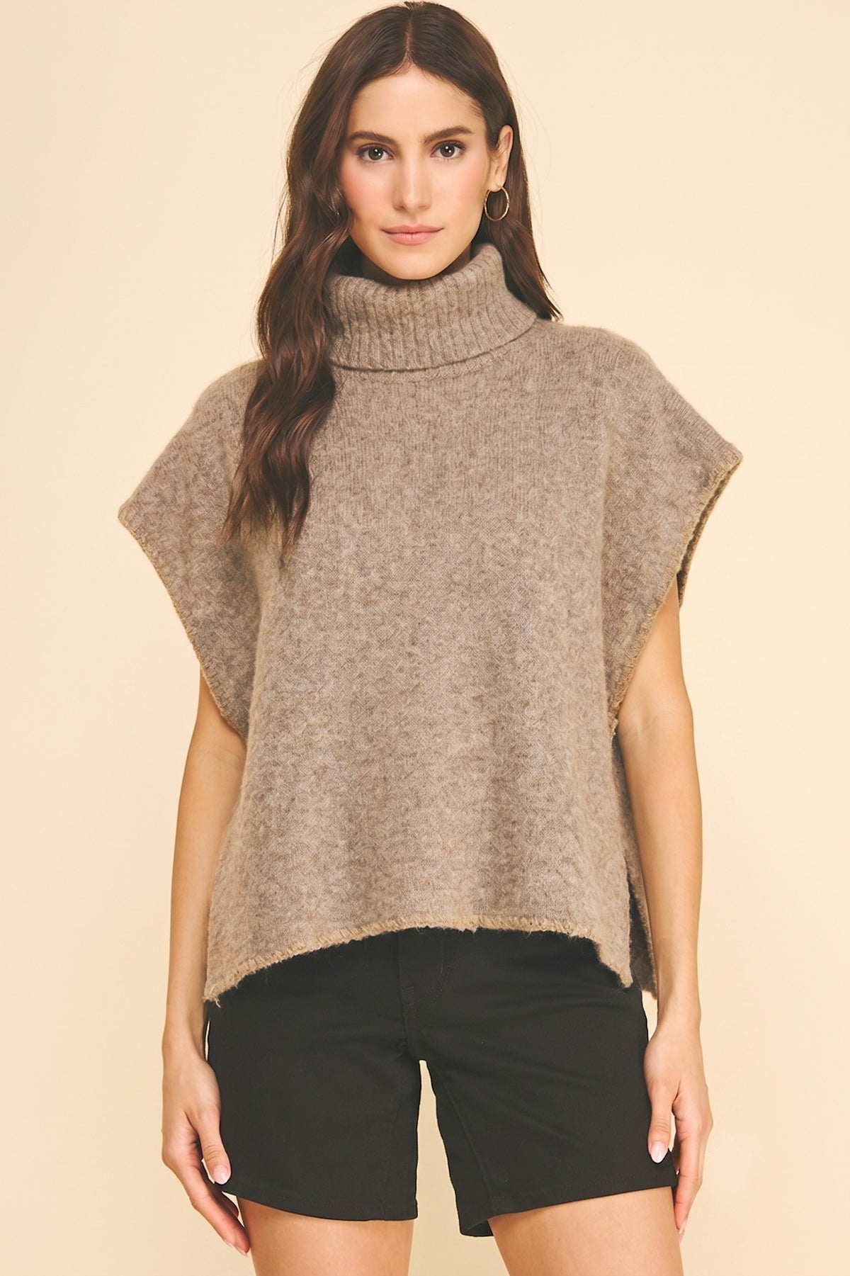 A woman modeling a chunky knit turtleneck poncho sweater in a heathered taupe-brown color, paired with black shorts, against a solid beige background.
