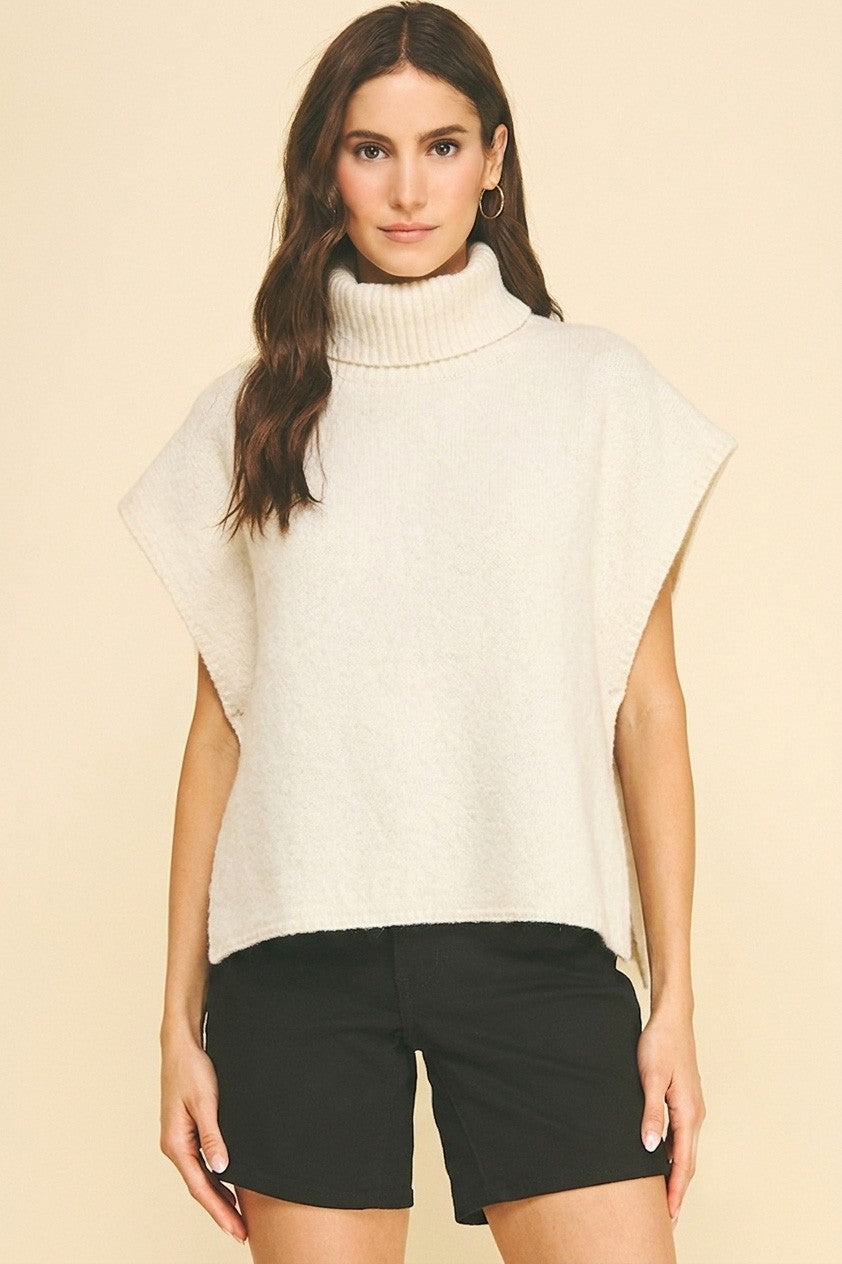 A woman modeling a cream-colored, sleeveless mock-neck knit top with wide draped shoulders, paired with black shorts, against a solid beige background.
