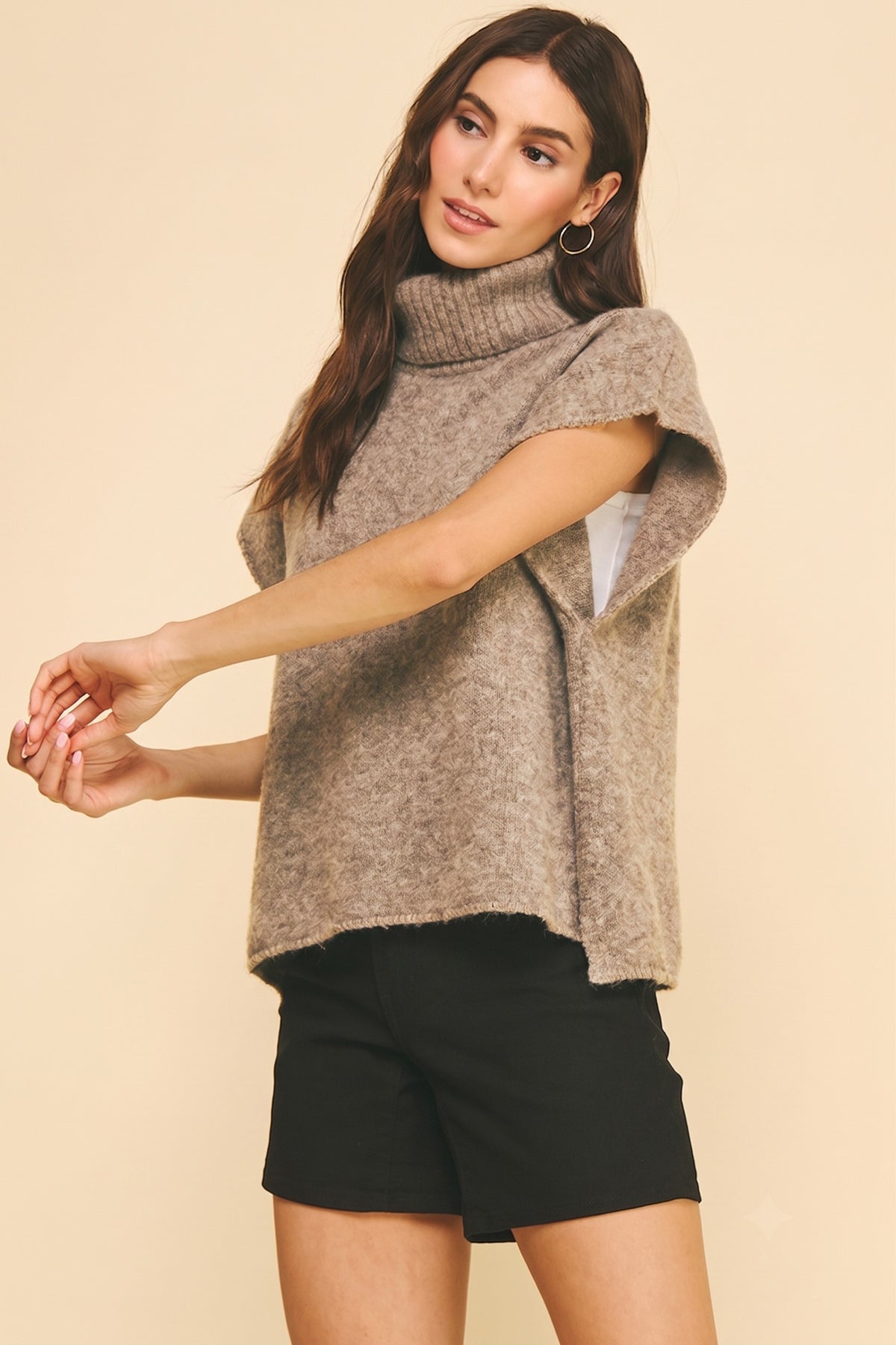 A woman modeling a fuzzy taupe-brown short-sleeve mock neck sweater with black shorts against a solid beige background.
