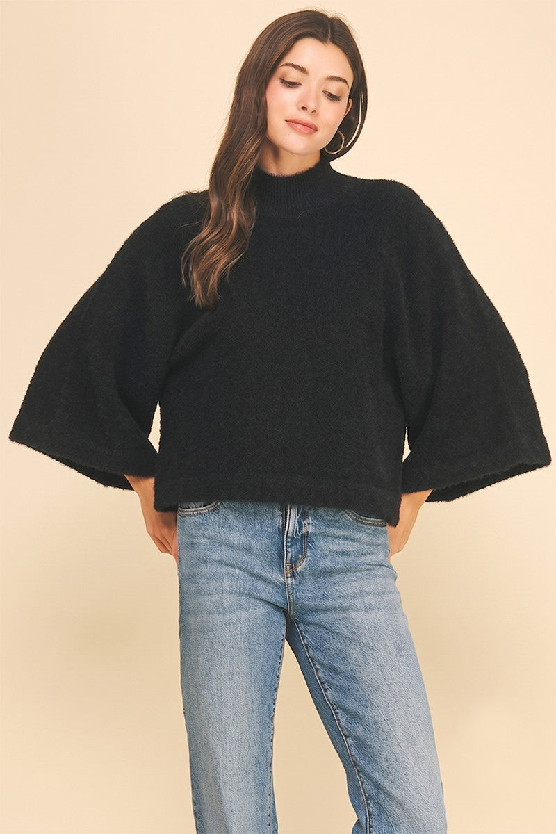 A woman wearing a black fuzzy knit mock-neck sweater with wide bell sleeves and light-wash blue jeans, standing against a solid beige studio background.