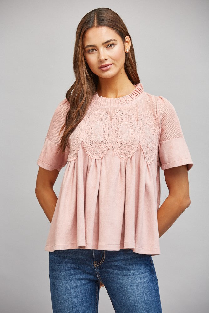 A woman wearing a dusty pink short-sleeve peplum top with intricate lace applique across the chest, paired with blue jeans, photographed against a neutral gray studio background.
