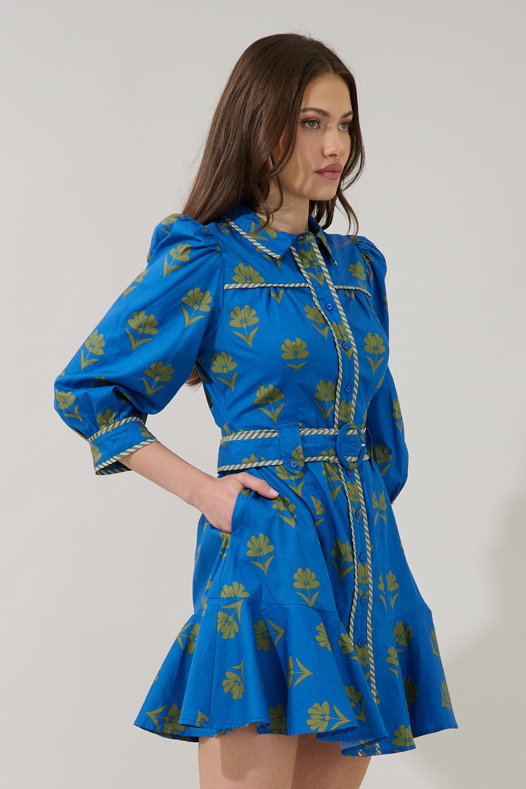 A woman modeling a bright blue mini dress with a yellow floral print, featuring a high collar, puffed sleeves, and a matching waist tie with decorative trim, set against a plain light gray background.