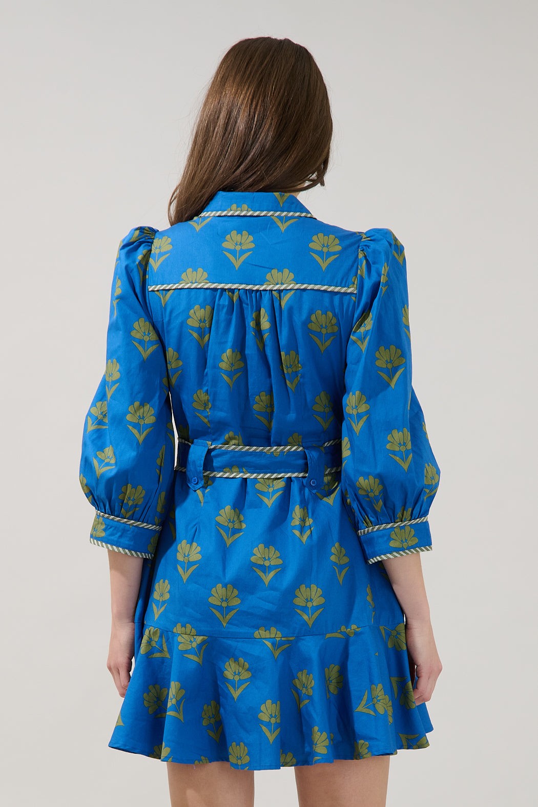 A woman viewed from the back wearing a bright blue mini dress with a gold floral block print and a rope-style tie belt at the waist, against a plain light gray studio background.