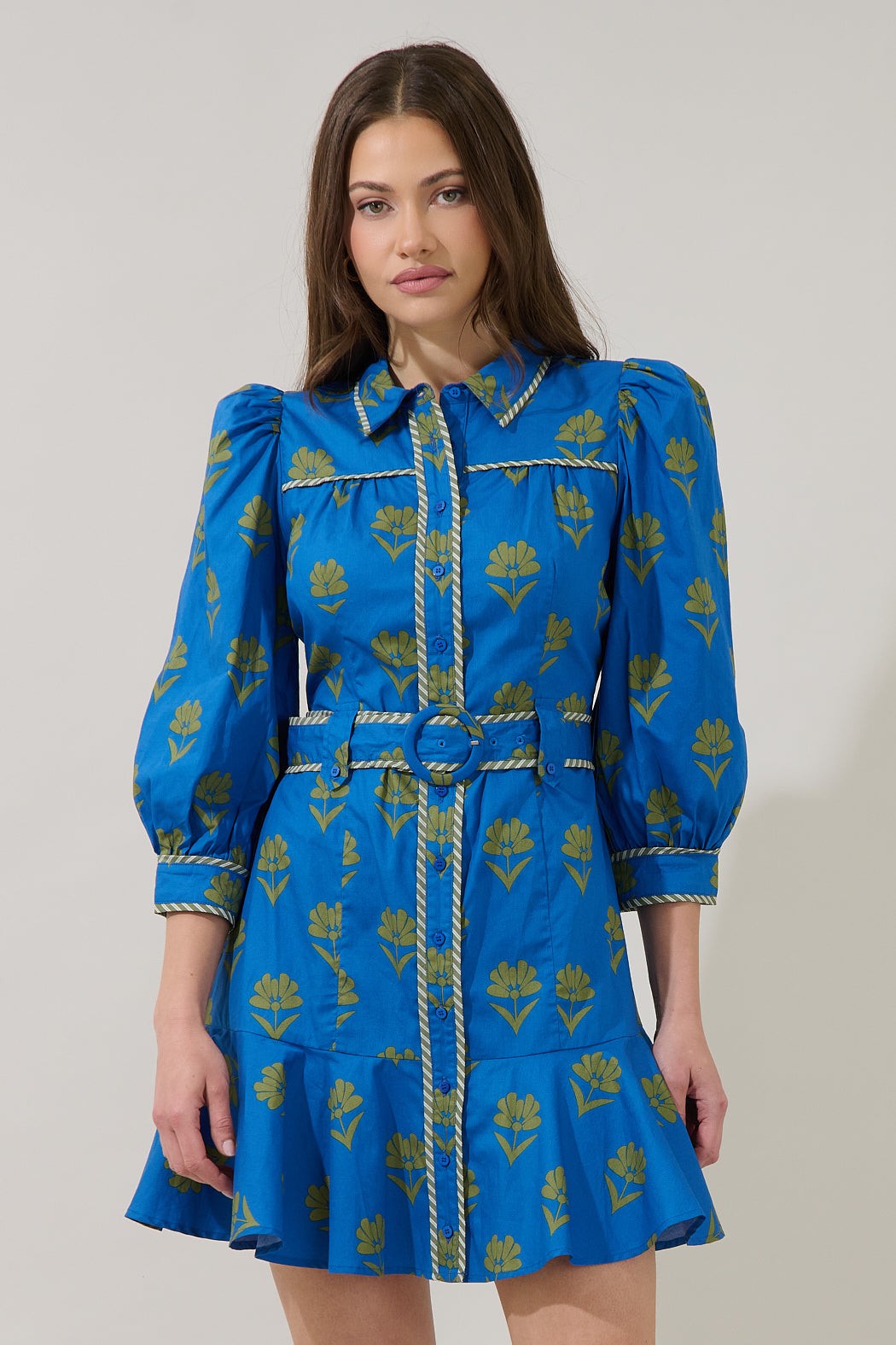 A woman modeling a bright blue mini shirt dress with a yellow floral print, featuring puffed sleeves, a matching belt, and decorative trim.