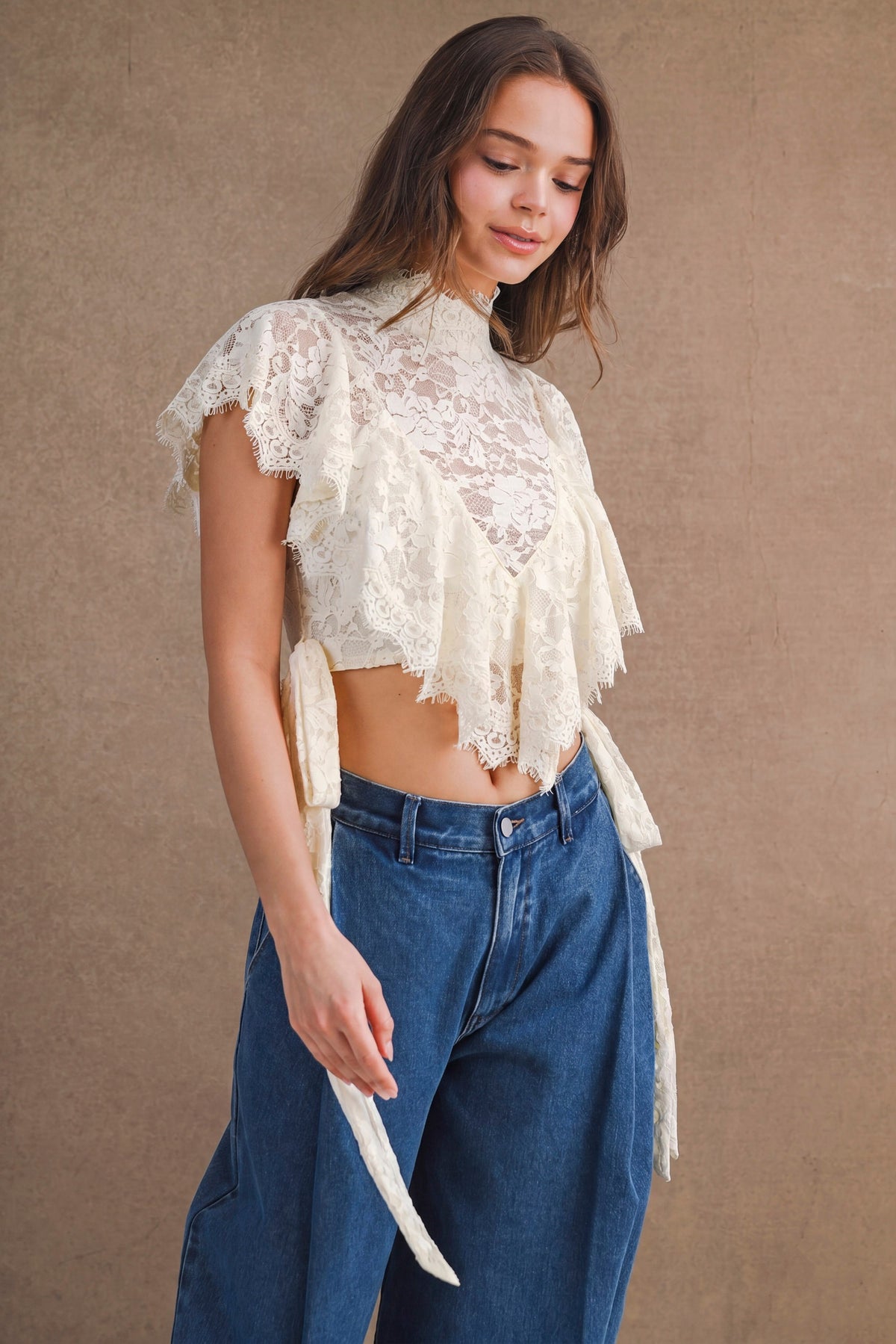 A woman wearing a cream lace cropped top with ruffle details and side ties, paired with high-waisted blue jeans, standing against a textured brown backdrop.