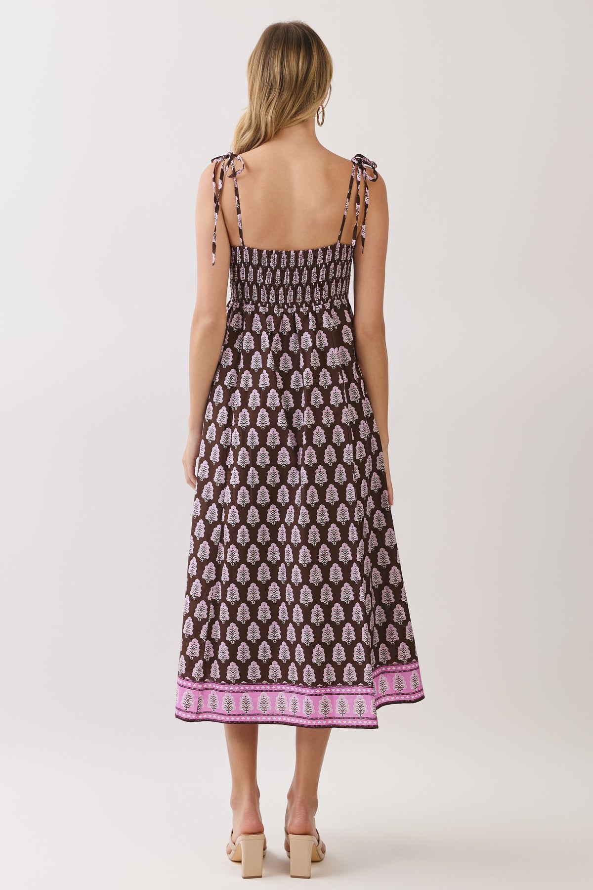 A woman modeling a sleeveless midi dress with thin shoulder ties, a brown base, and a repeating pink and white paisley-like print, finished with a pink patterned border at the hem, against a light gray studio background.