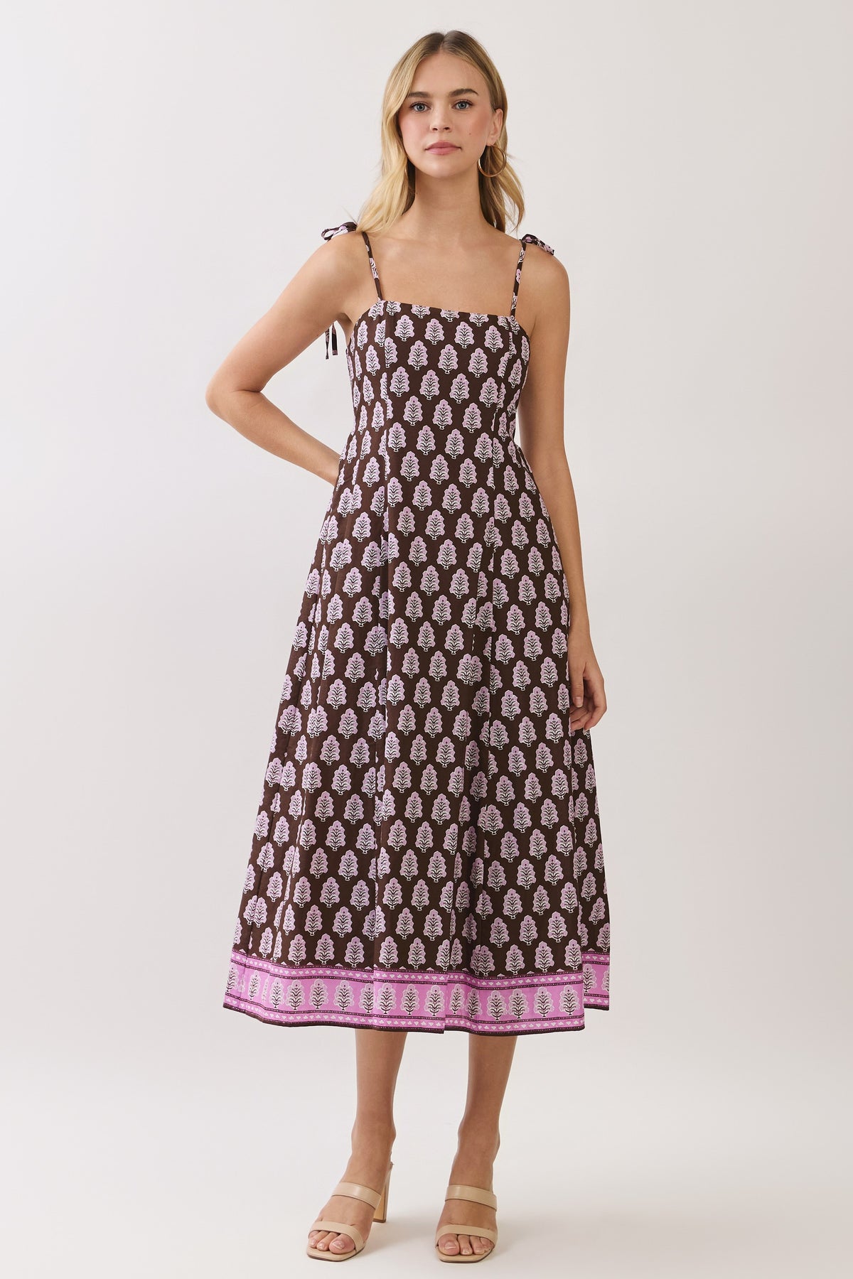 A woman modeling a sleeveless midi dress with thin shoulder ties, a brown base, and a repeating pink and white paisley-like print, finished with a pink patterned border at the hem, against a light gray studio background.