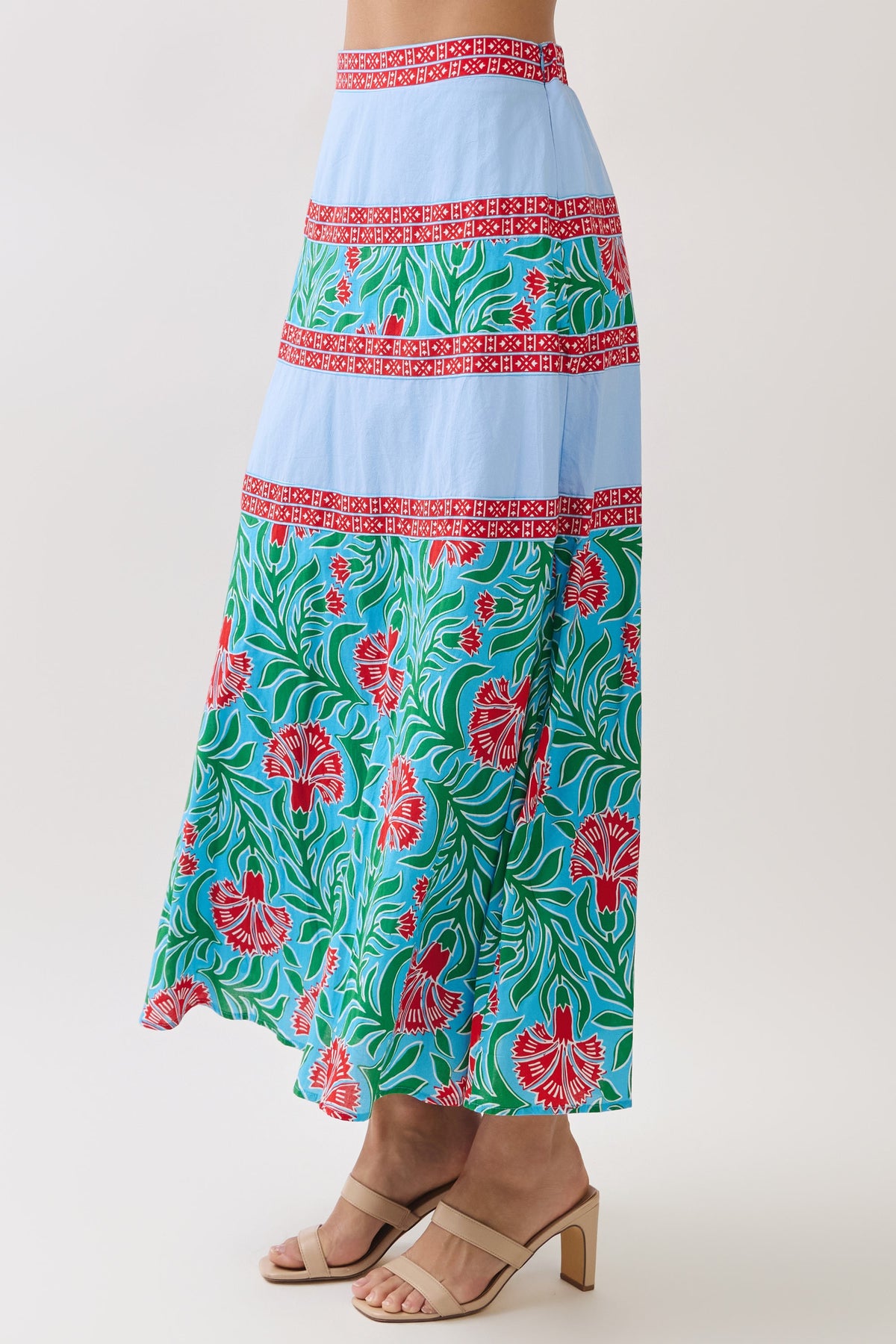 A model wearing a tiered maxi skirt with a light blue base, red patterned horizontal bands, and a green tropical floral print on the lower half, paired with beige heeled sandals against a light gray studio background.