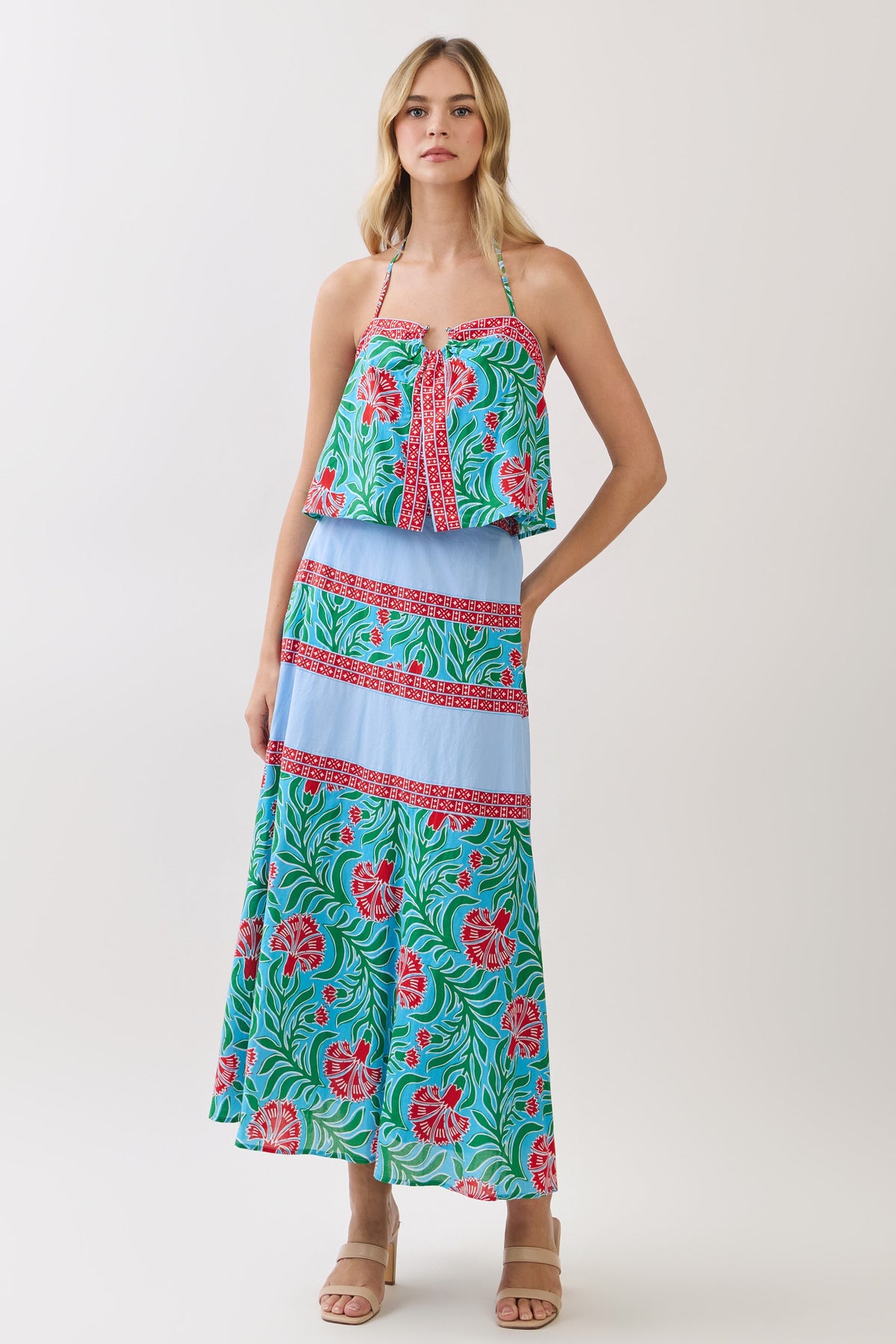 A woman modeling a halter-neck maxi dress with a split design: a red, green, and blue floral corset-style bodice and a light blue pleated skirt with matching patterned bands, against a plain light background.