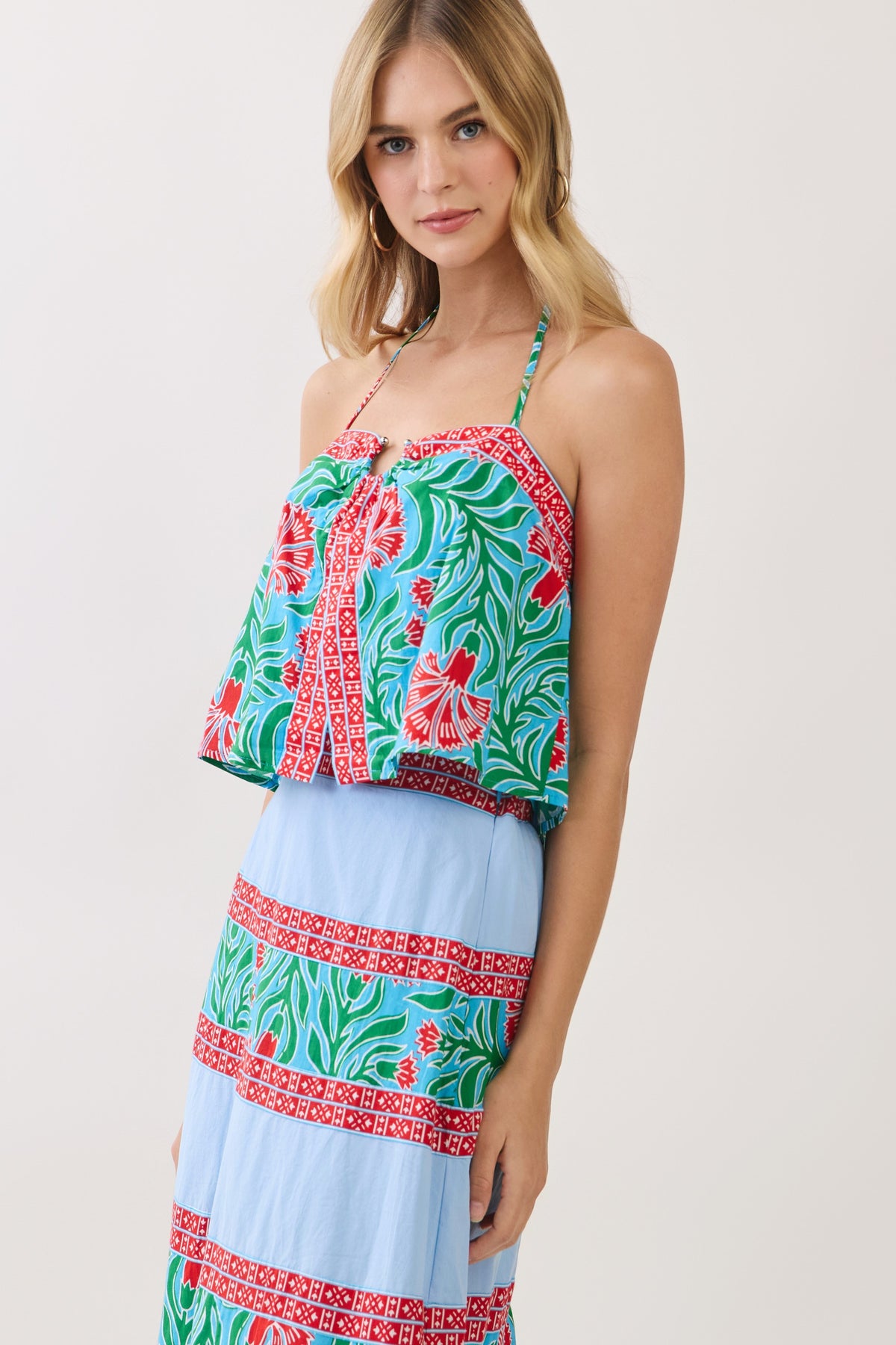 A woman modeling a two-piece dress set featuring a green tropical floral print crop top and a light blue tiered skirt with red patterned bands, set against a plain light gray studio background.