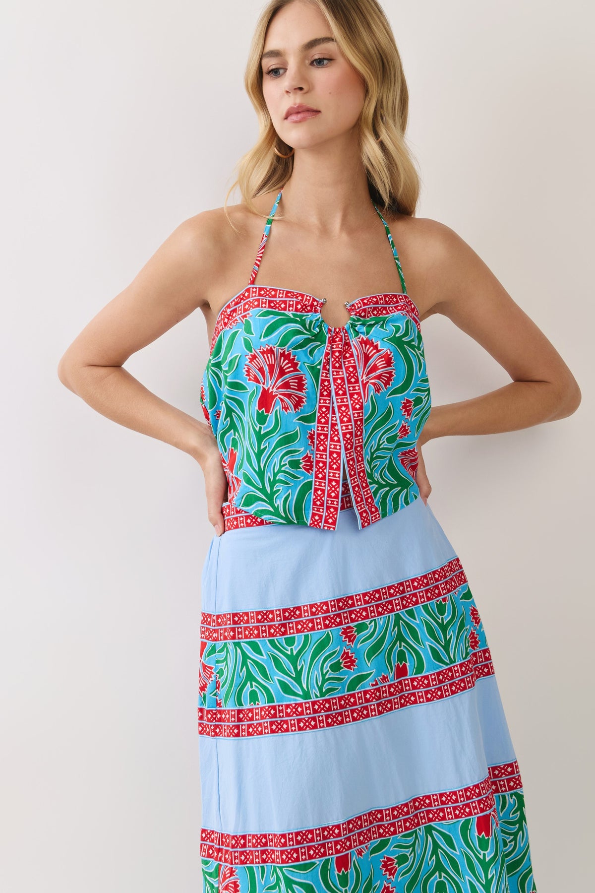 A woman modeling a halter-neck maxi dress with a split design: a red, green, and blue floral corset-style bodice and a light blue pleated skirt with matching patterned bands, against a plain light background.