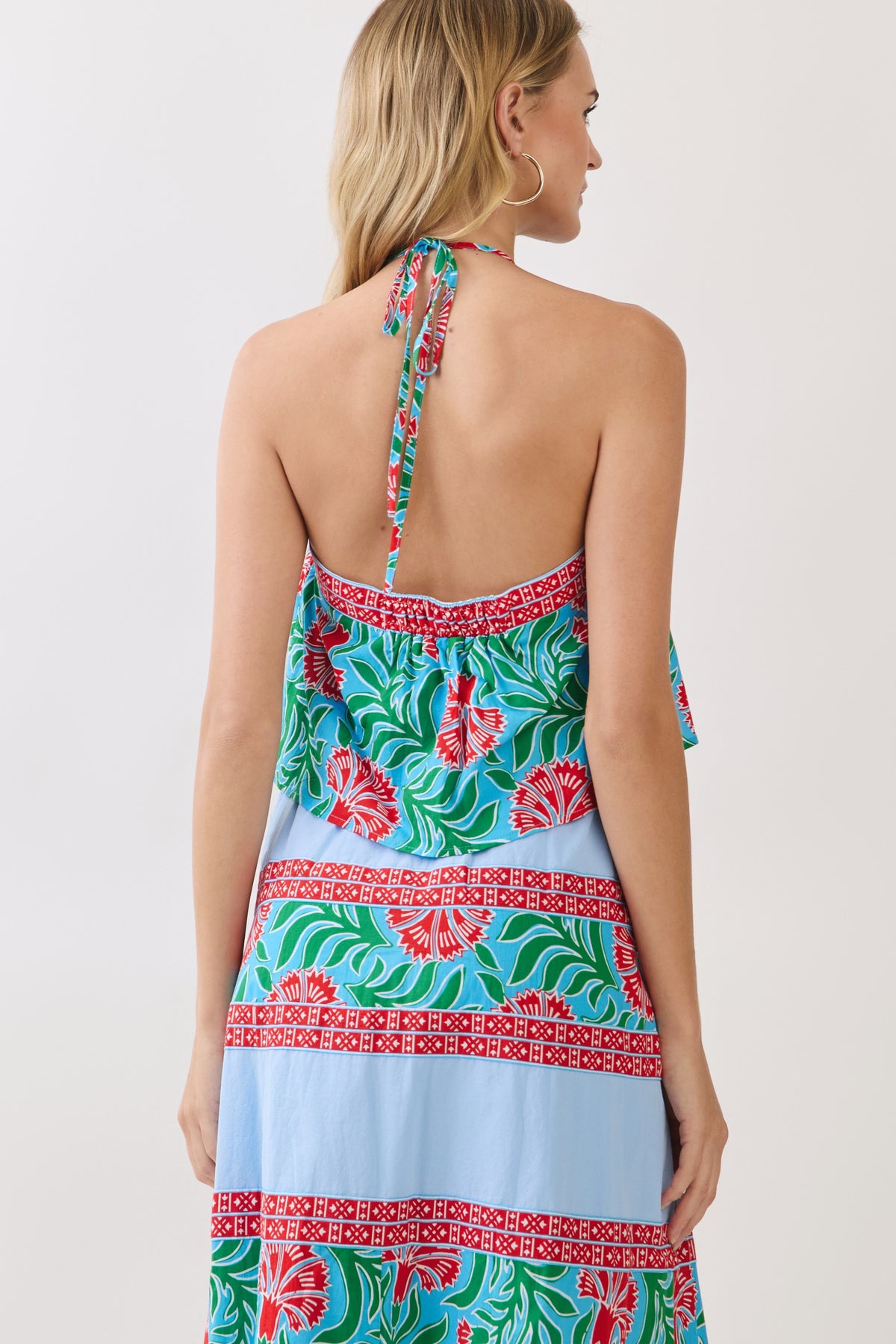A woman shown from the back wearing a halter-neck dress with a colorful tropical floral print and light blue horizontal stripes, photographed against a plain light gray studio background.