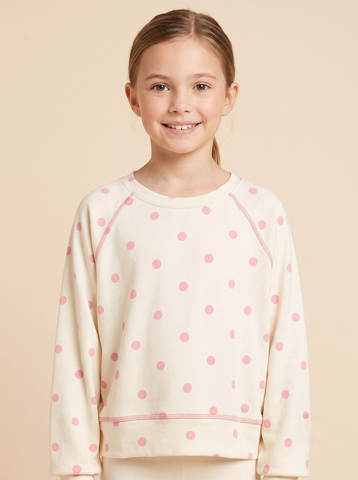 A young girl smiling and wearing a cream-colored long-sleeve sweatshirt with pink polka dots and matching pink trim, set against a solid beige background.