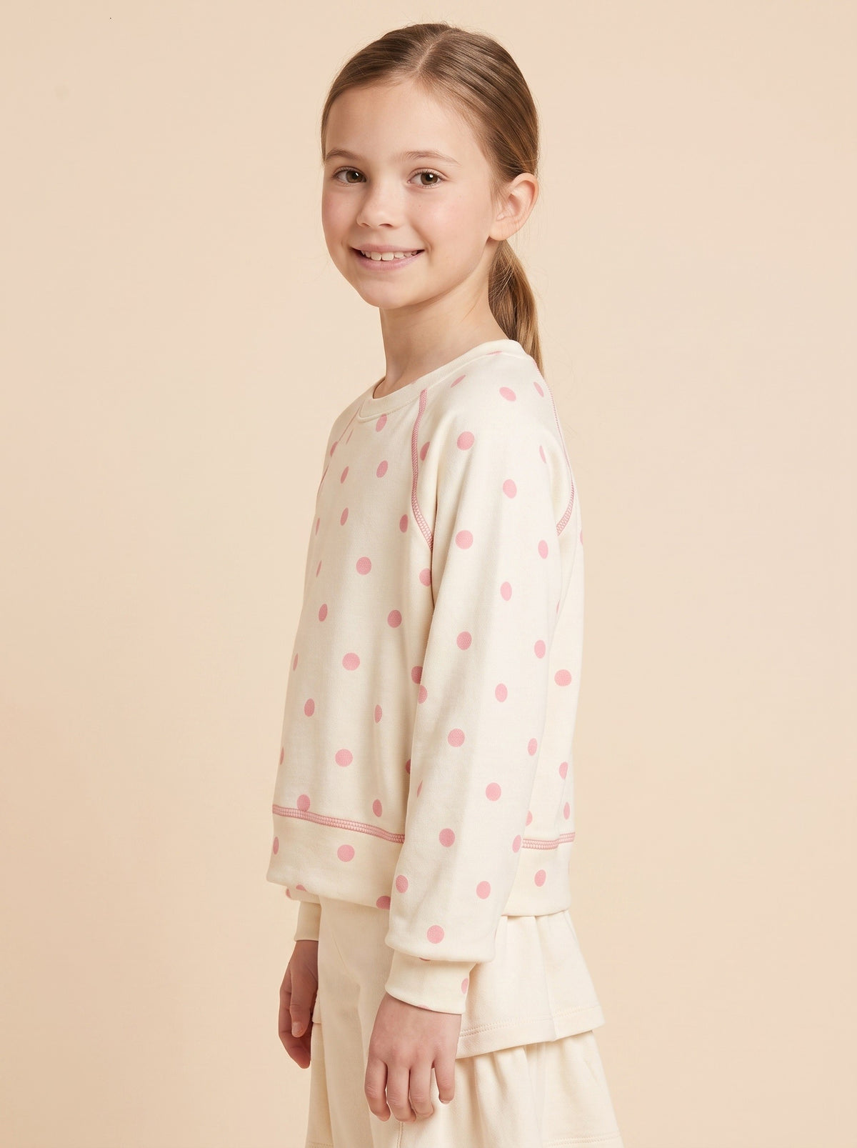 A young girl with her hair in a ponytail is shown from the side, wearing a cream-colored long-sleeve sweatshirt with pink polka dots and matching cream pants, set against a solid light beige background.