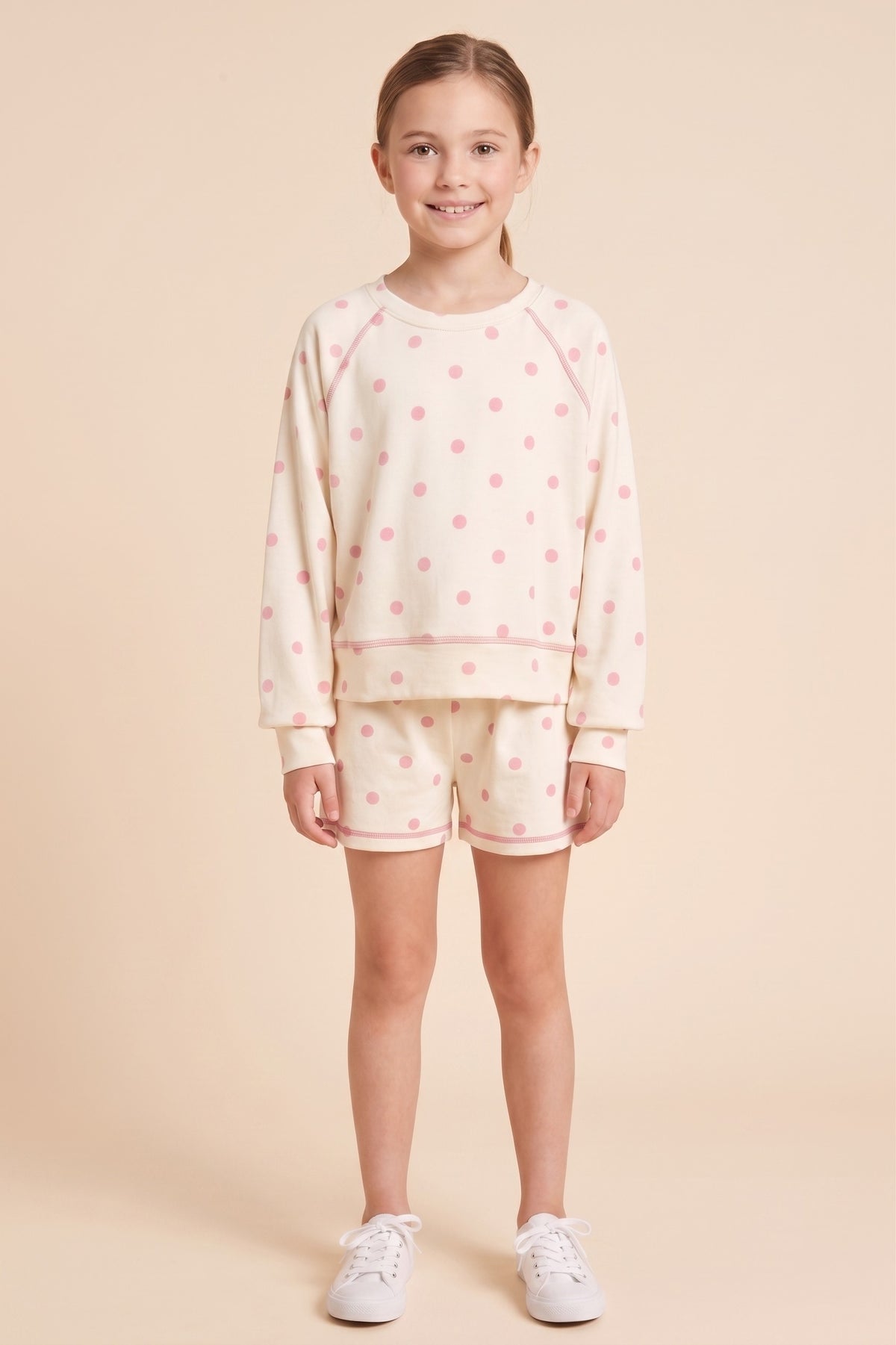 A young girl stands against a solid beige studio background wearing a matching cream-colored two-piece lounge set with pink polka dots, consisting of a long-sleeve sweatshirt and shorts, paired with white sneakers.