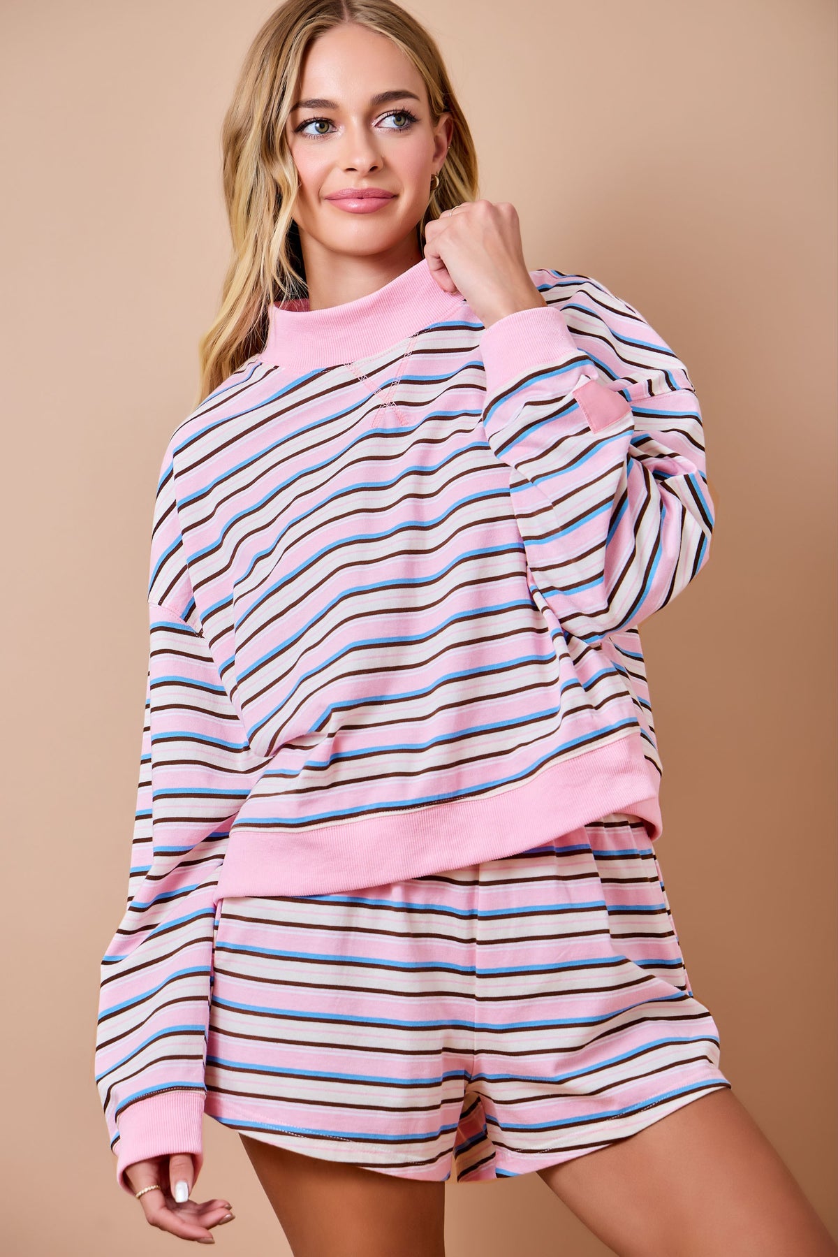 A woman modeling a matching two-piece lounge set featuring a pink mock neck sweatshirt and shorts with horizontal multicolor stripes in pink, blue, and black on a beige background.