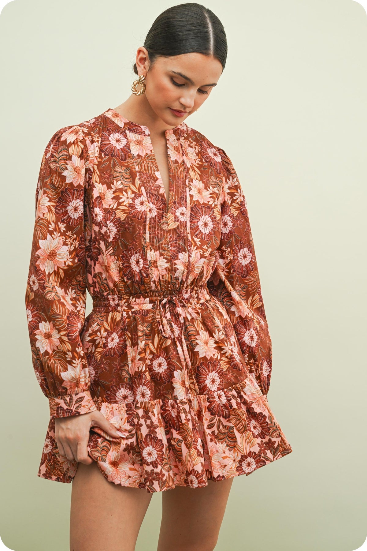 A woman wearing a long-sleeve mini dress with a brown, rust, and pink floral pattern, standing against a solid light beige background.