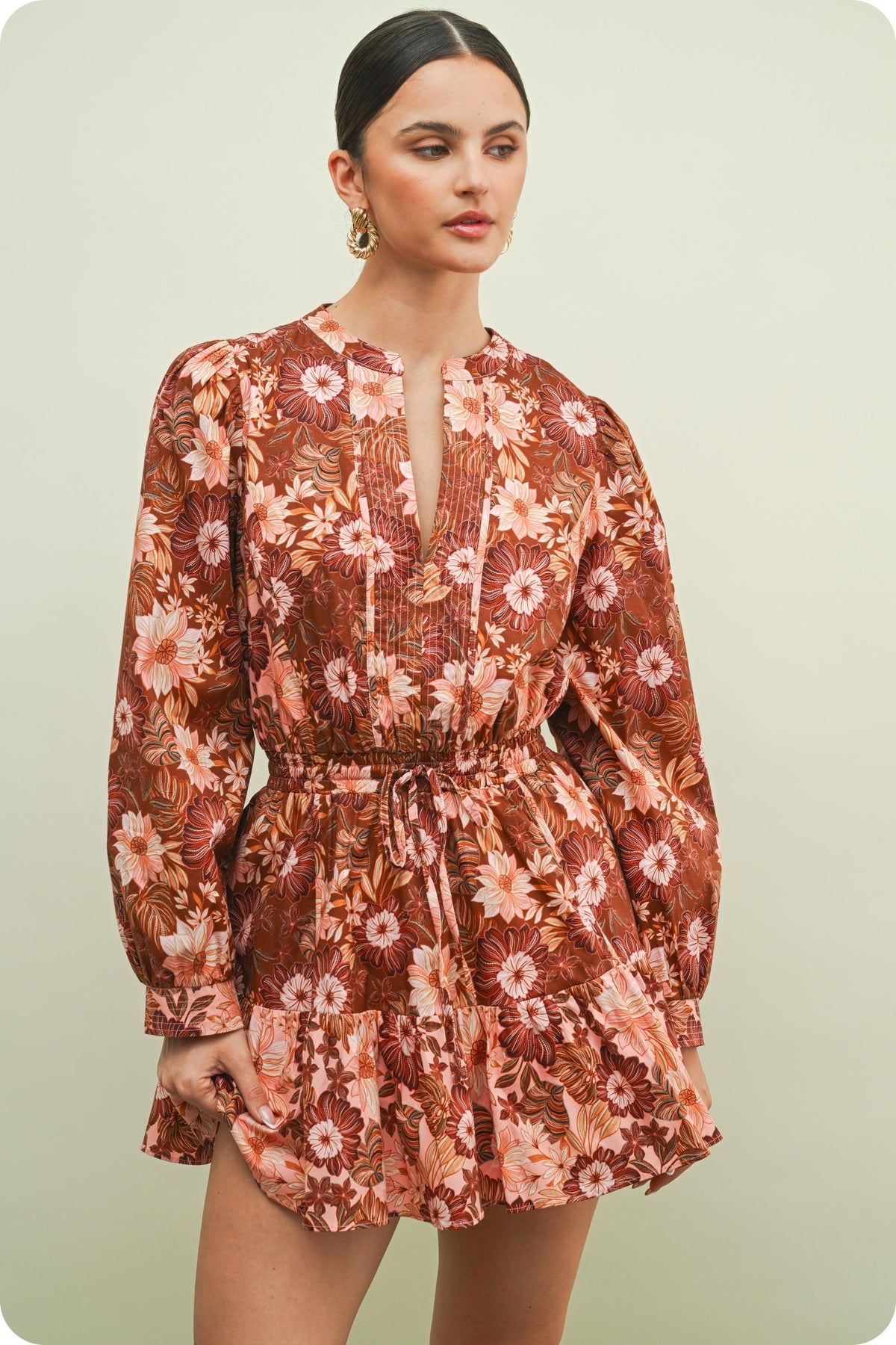 A woman modeling a long-sleeve mini dress with a brown, rust, and pink floral pattern against a solid light green background.