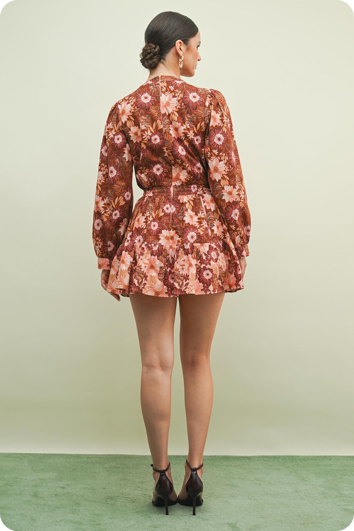 A woman is shown from the back wearing a long-sleeve mini dress with a brown, rust, and peach floral pattern. She is standing against a light beige studio background with a green floor.
