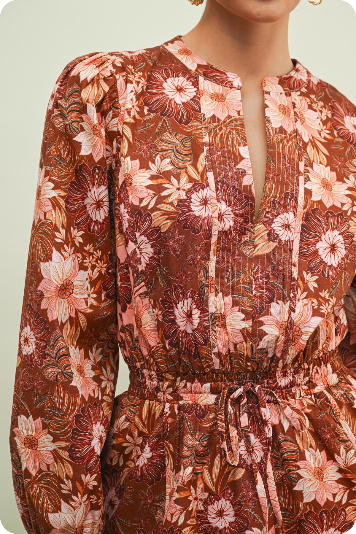 A close-up of a woman wearing a long-sleeve blouse with a brown, rust, and pink floral pattern, featuring a split neckline and a drawstring waist.