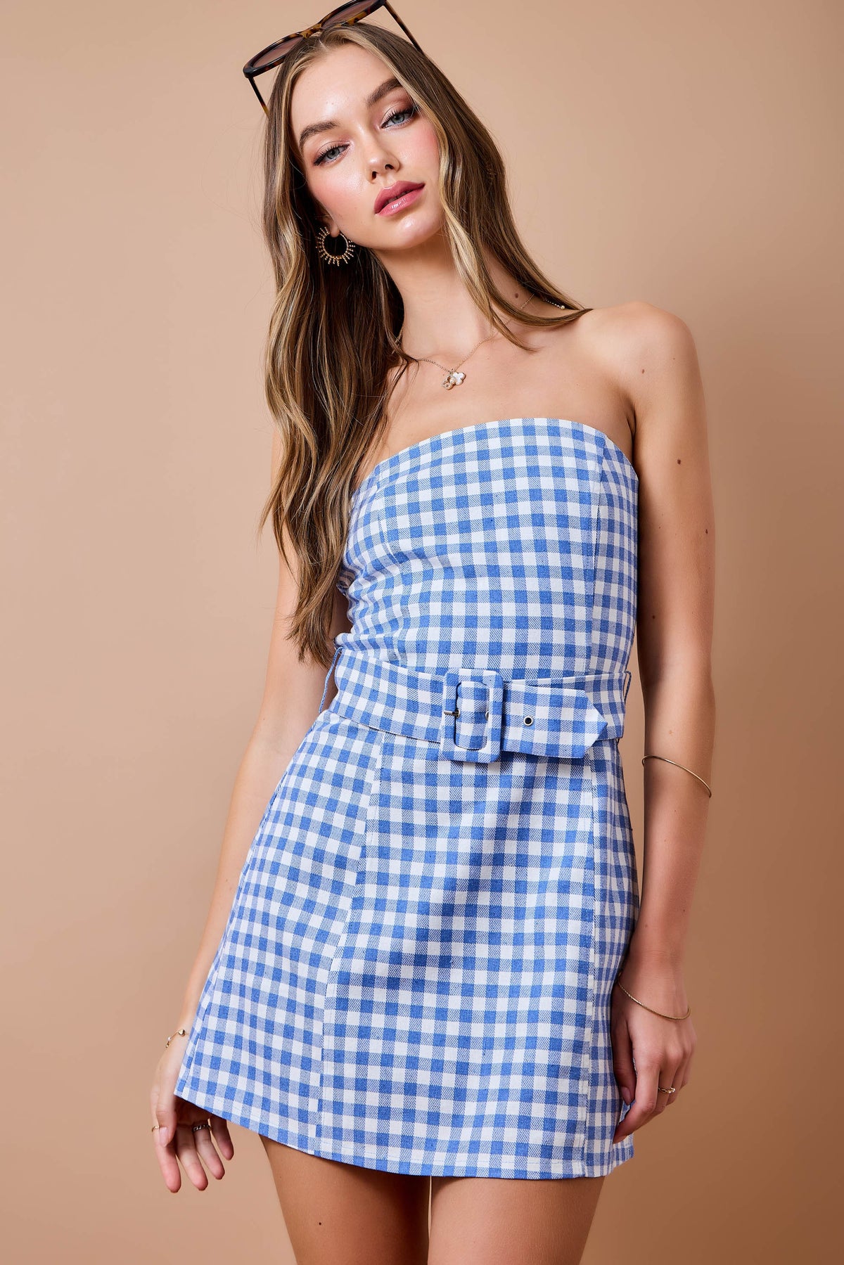 A woman modeling a strapless blue and white gingham mini dress with a matching fabric belt and large square buckle, posed against a solid beige studio background.
