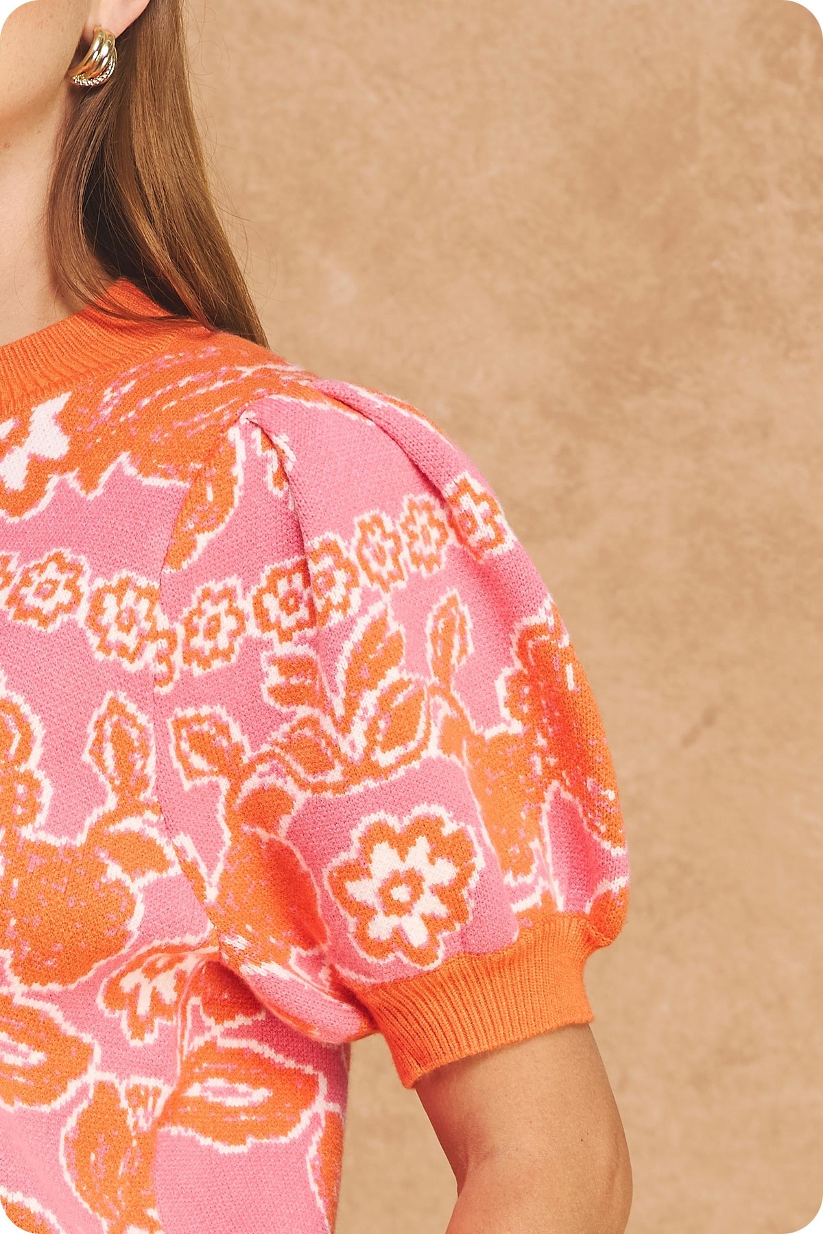 A close-up of a model wearing a pink knit sweater with a bold orange floral and abstract pattern, featuring puff sleeves and ribbed cuffs.