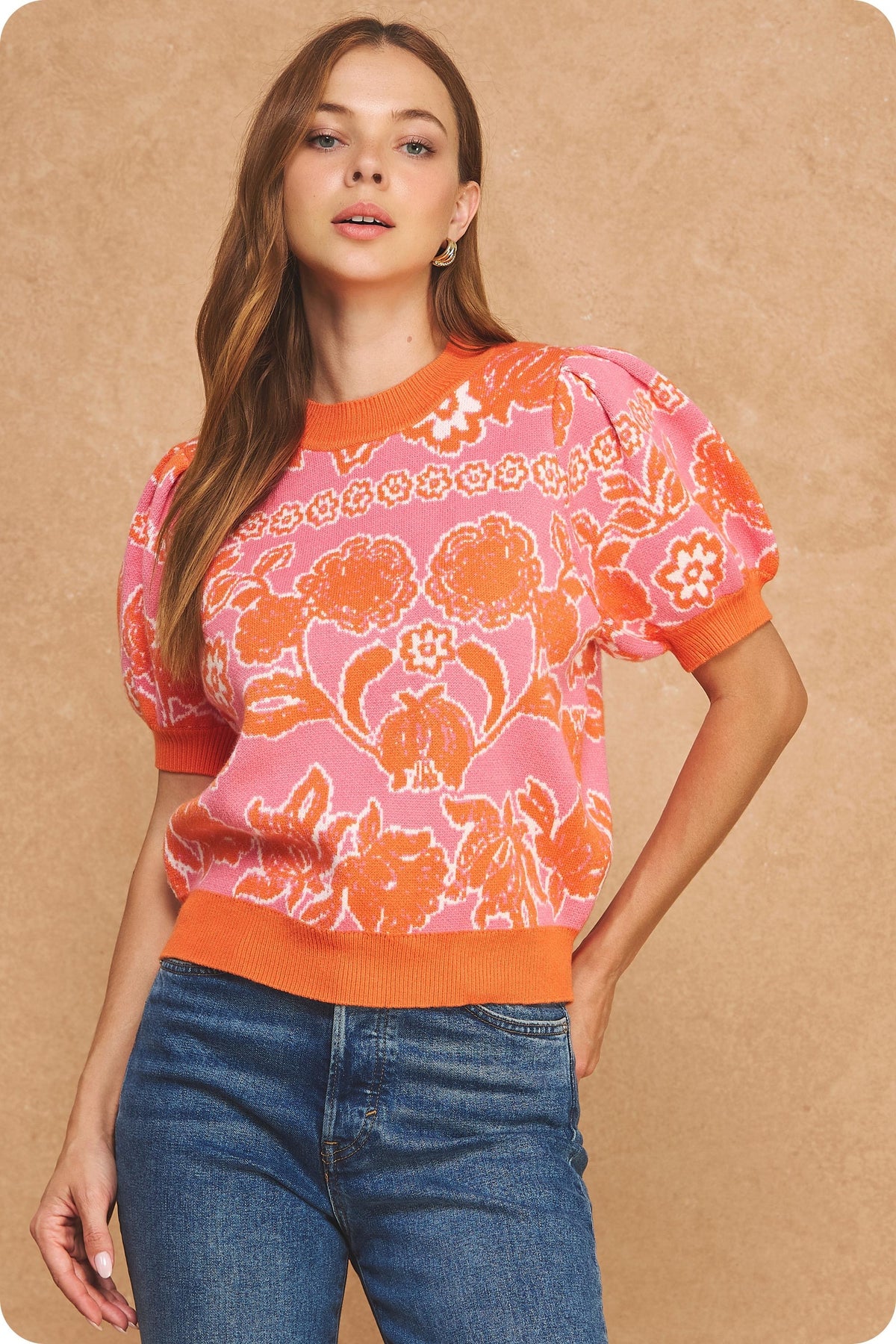 A woman modeling a short-sleeve knit sweater with a pink and orange floral pattern, paired with blue jeans, against a textured beige background.