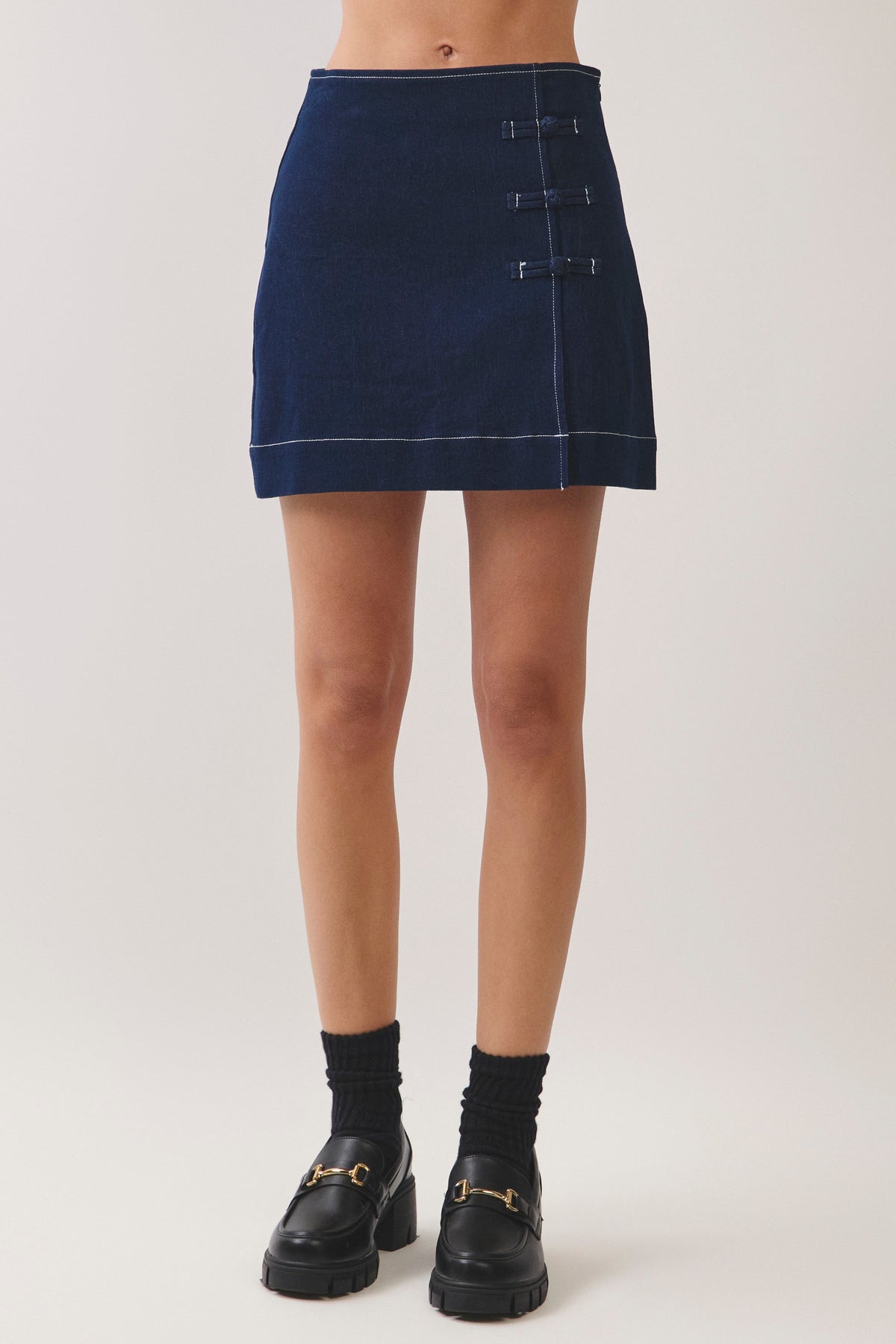 A model from the waist down wearing a dark blue denim mini skirt with decorative metal button details on the side, paired with black socks and black leather loafers with gold hardware, against a light gray studio background.