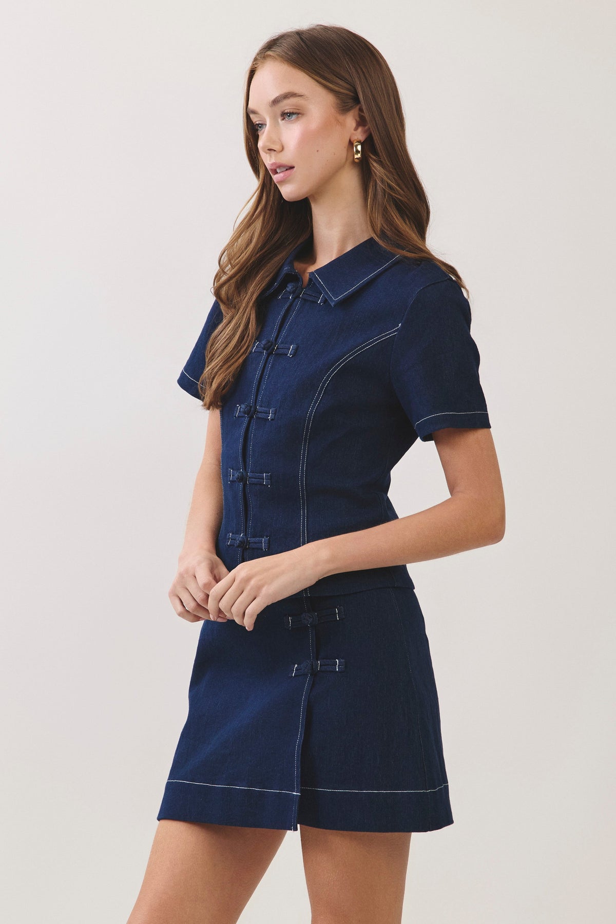 A woman modeling a dark navy blue denim short-sleeve shirt and matching mini skirt set with contrast white stitching and traditional Chinese-style frog closures, set against a plain light gray studio background.