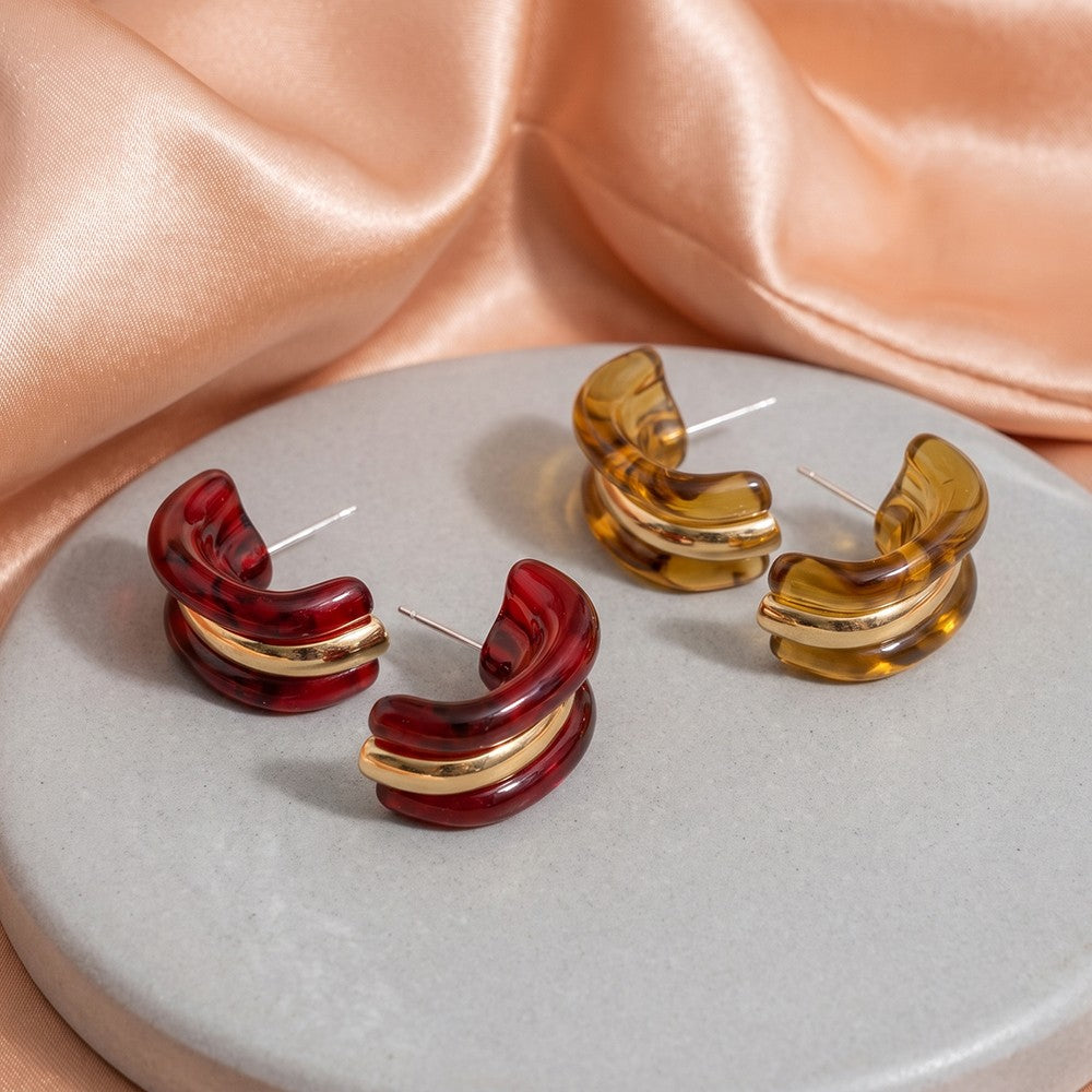 Three pairs of curved hoop earrings made of translucent amber and deep red resin with polished gold-tone inner bands, arranged on a round gray display plate with peach satin fabric in the background.