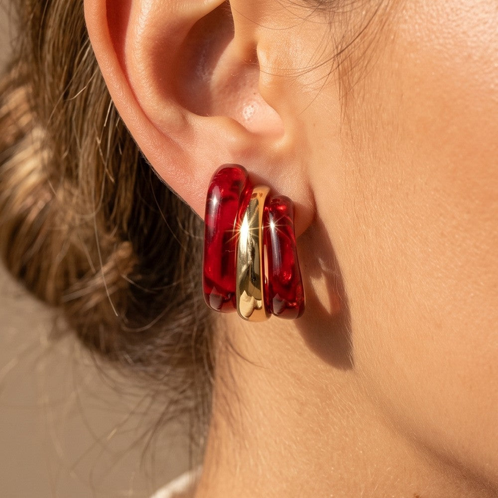 A close-up of a person's ear wearing a pair of chunky, stacked hoop earrings made of translucent deep red resin with a shiny gold center accent.