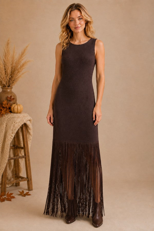 A woman models a sleeveless, bodycon maxi dress in dark brown suede-like fabric with a long fringe hem, standing against a warm beige studio backdrop with decorative autumn props on the left.
