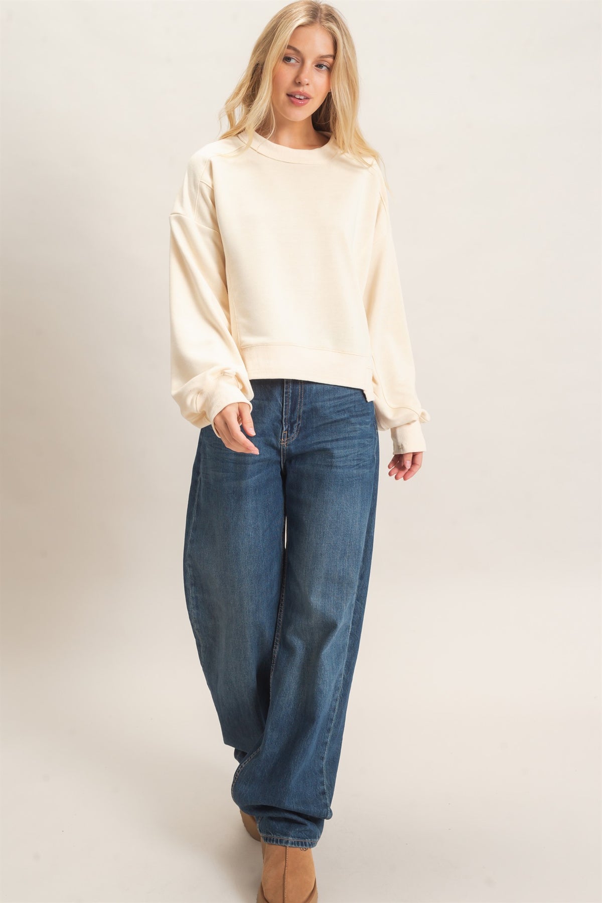 A woman modeling a cream-colored long-sleeve crewneck sweatshirt and wide-leg blue jeans against a plain beige studio background.