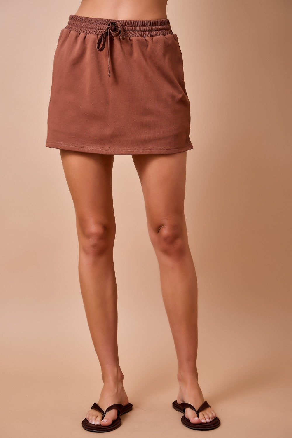 A person wearing a brown drawstring mini skirt with a relaxed, straight silhouette, paired with black flip-flop sandals, shown against a solid beige studio background.