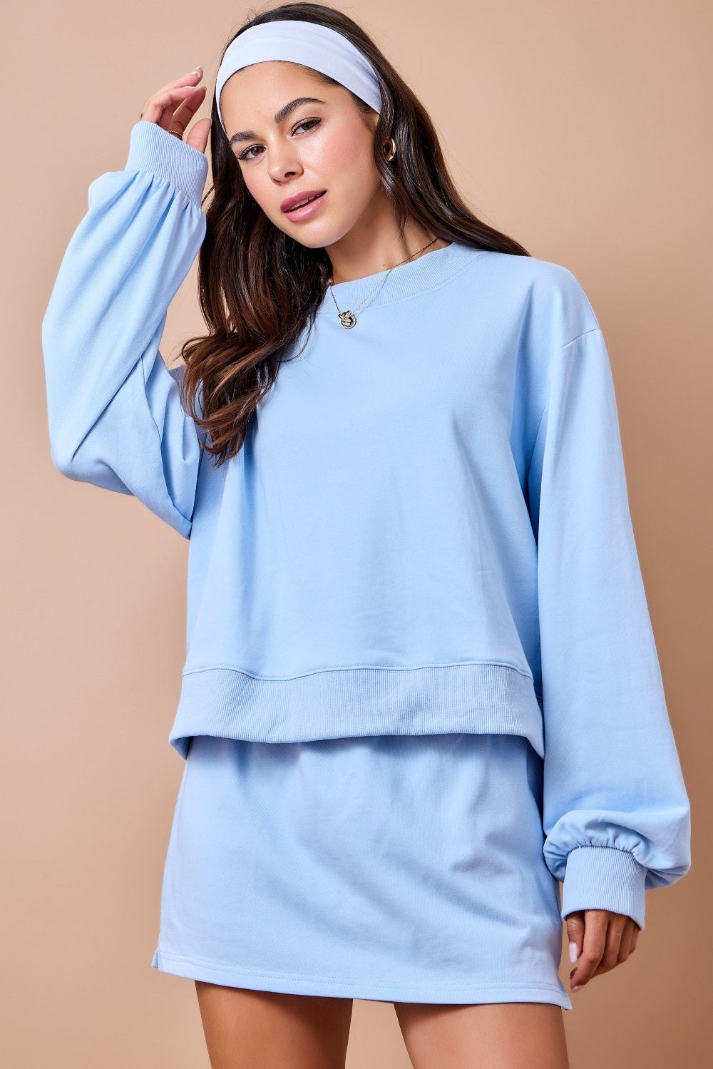 A woman modeling a light blue sweatshirt and matching mini skirt set against a solid beige background. She is wearing a matching light blue headband.