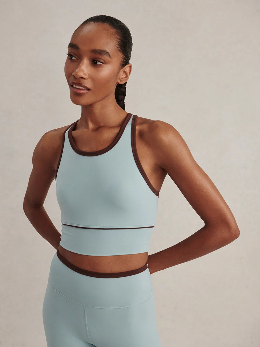 A woman modeling a light blue athletic crop top and matching leggings with dark brown trim, set against a neutral beige background.
