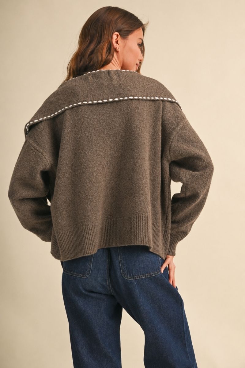A woman shown from the back wearing an oversized brown knit sweater with a white dotted trim detail across the shoulders, paired with dark blue jeans, against a plain beige background.