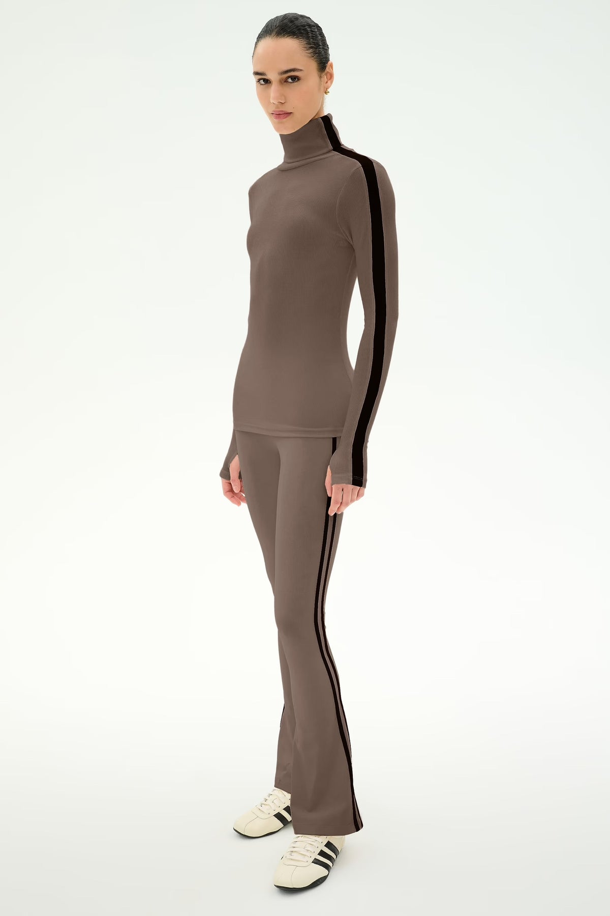 A model wearing a matching taupe-brown two-piece tracksuit with black side stripes, consisting of a mock-neck long-sleeve top and matching flared pants, paired with white sneakers.