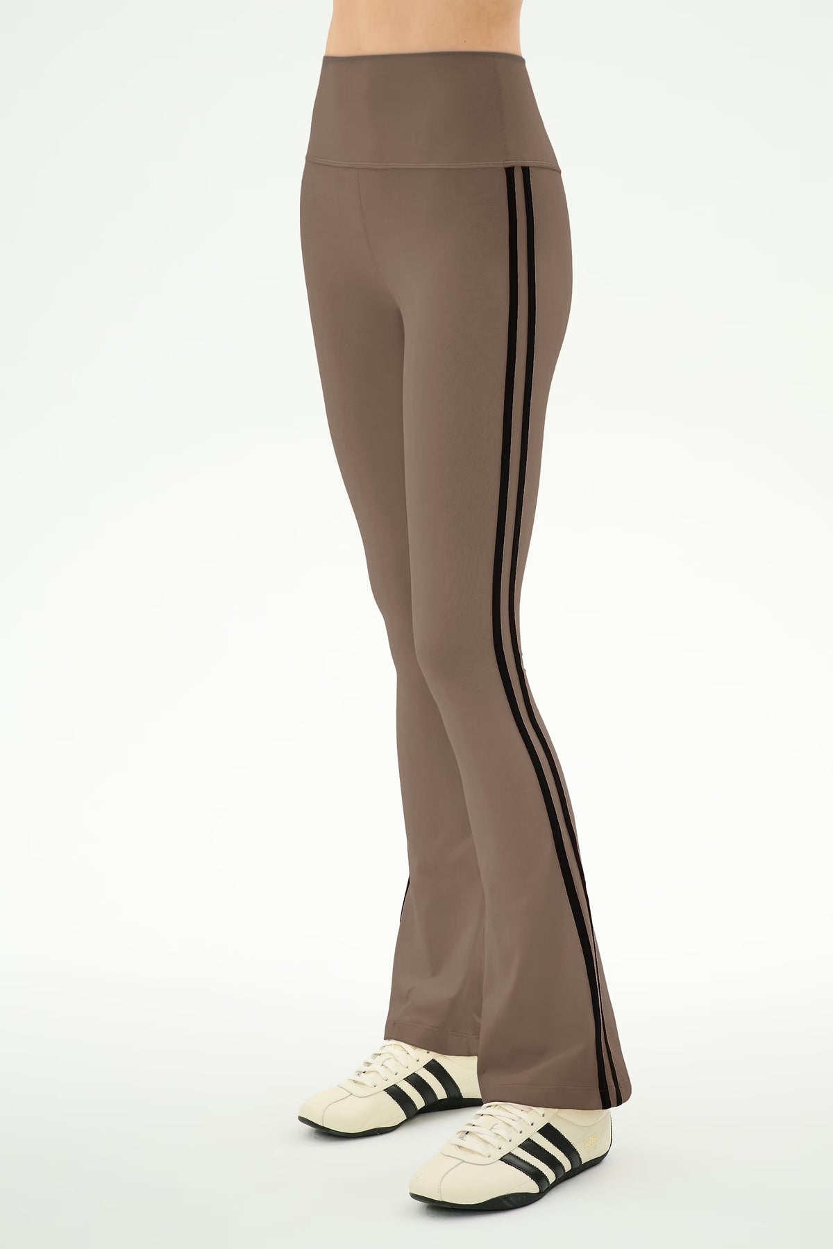 A person wearing high-waisted taupe-brown flared leggings with black side stripes, paired with cream sneakers with black stripes, shown from the waist down against a plain white studio background.