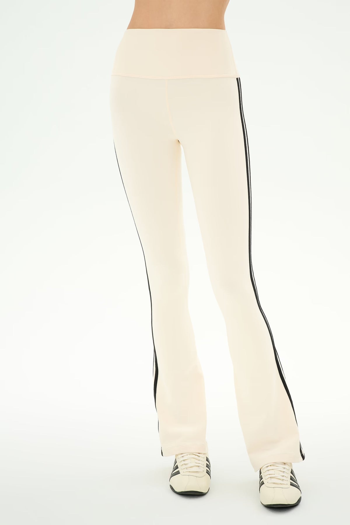 Cream-colored flared leggings with black side stripes, shown on a model from the waist down wearing white sneakers against a white studio background.