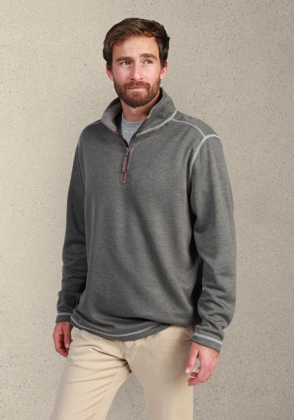 A man wearing a heather gray quarter-zip pullover sweater with contrasting light gray piping on the shoulders and cuffs, paired with beige pants, standing against a textured gray wall.