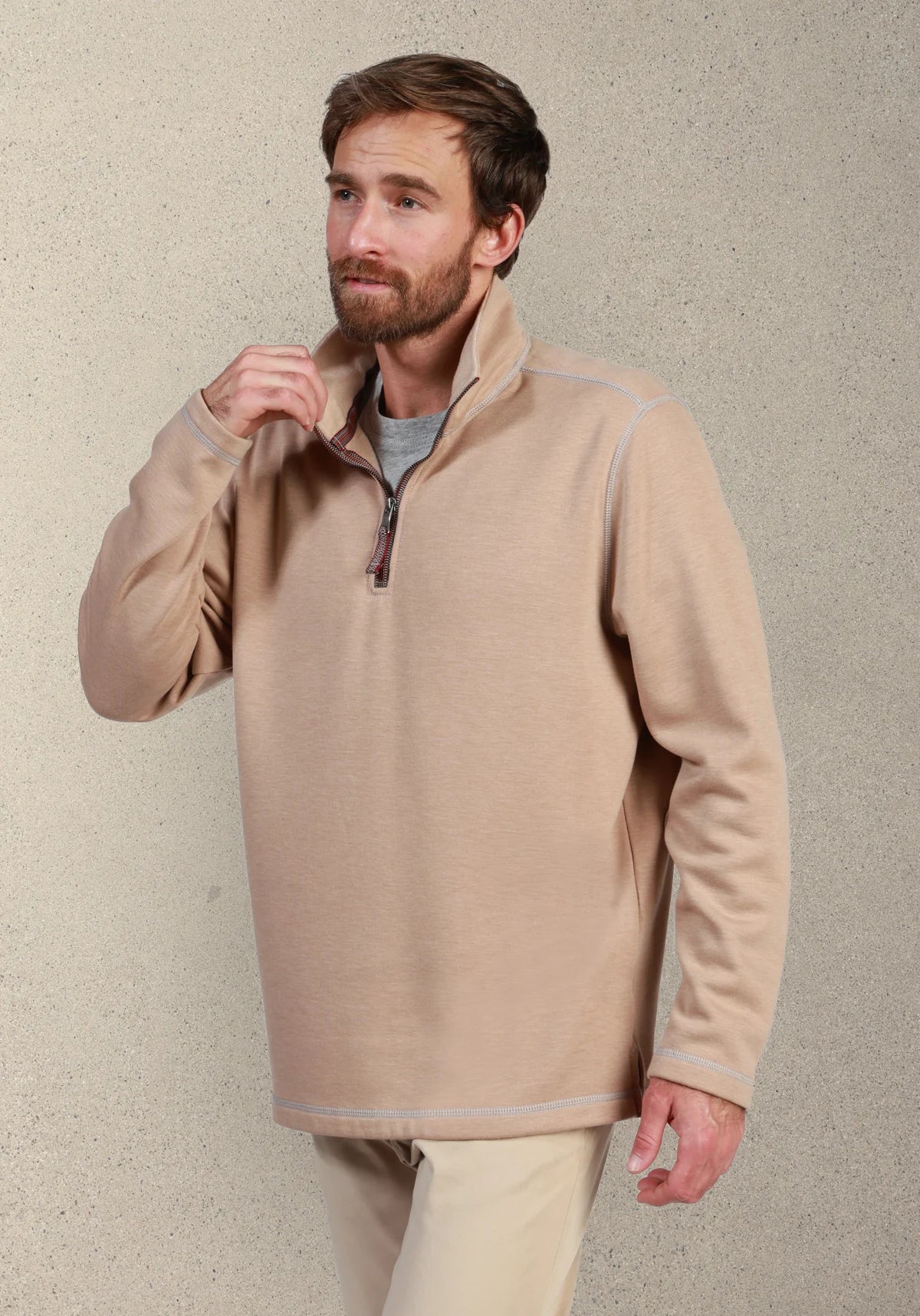 A man wearing a light beige quarter-zip fleece pullover sweater over a gray t-shirt and beige trousers, standing against a textured light gray wall.