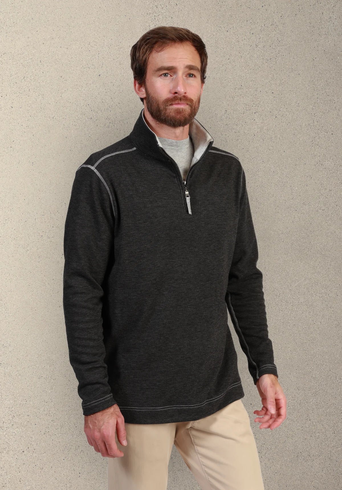 A man wearing a dark charcoal gray quarter-zip pullover sweater with light gray piping details, layered over a light gray t-shirt and paired with beige pants, standing against a textured beige wall.