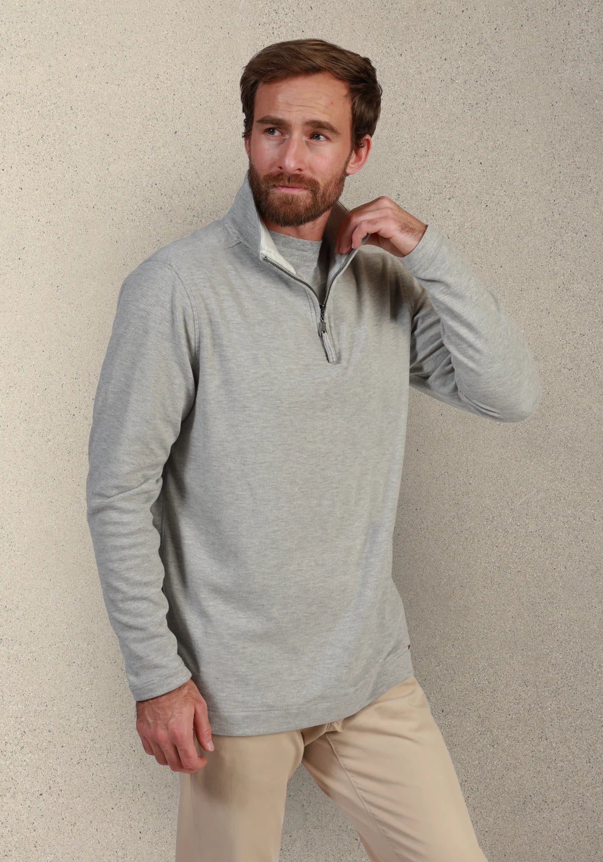 A man wearing a light gray quarter-zip long-sleeve pullover sweater and beige pants, standing against a textured beige wall.