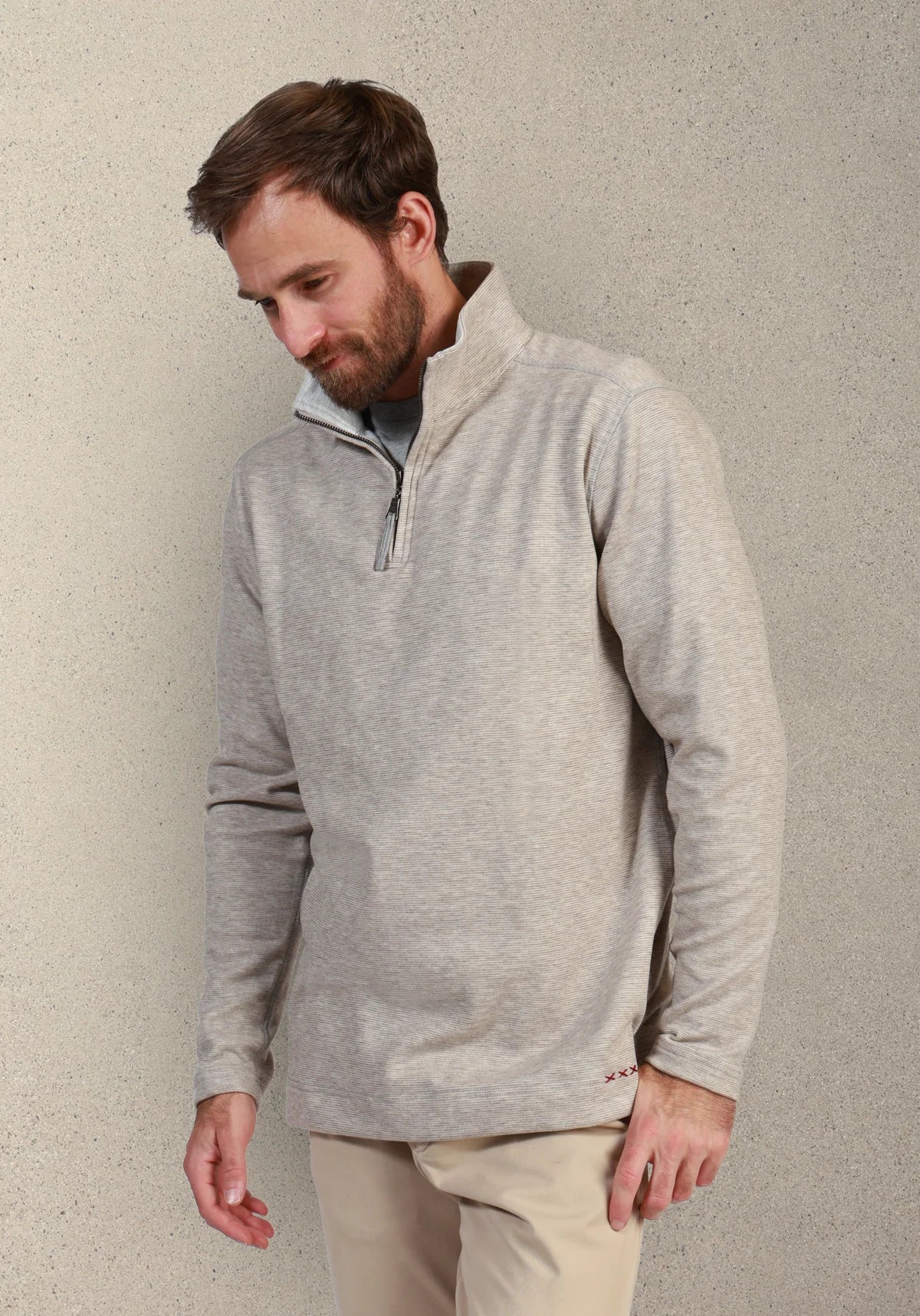 A man wearing a light gray heathered quarter-zip pullover sweater and beige trousers, standing against a textured light gray wall.