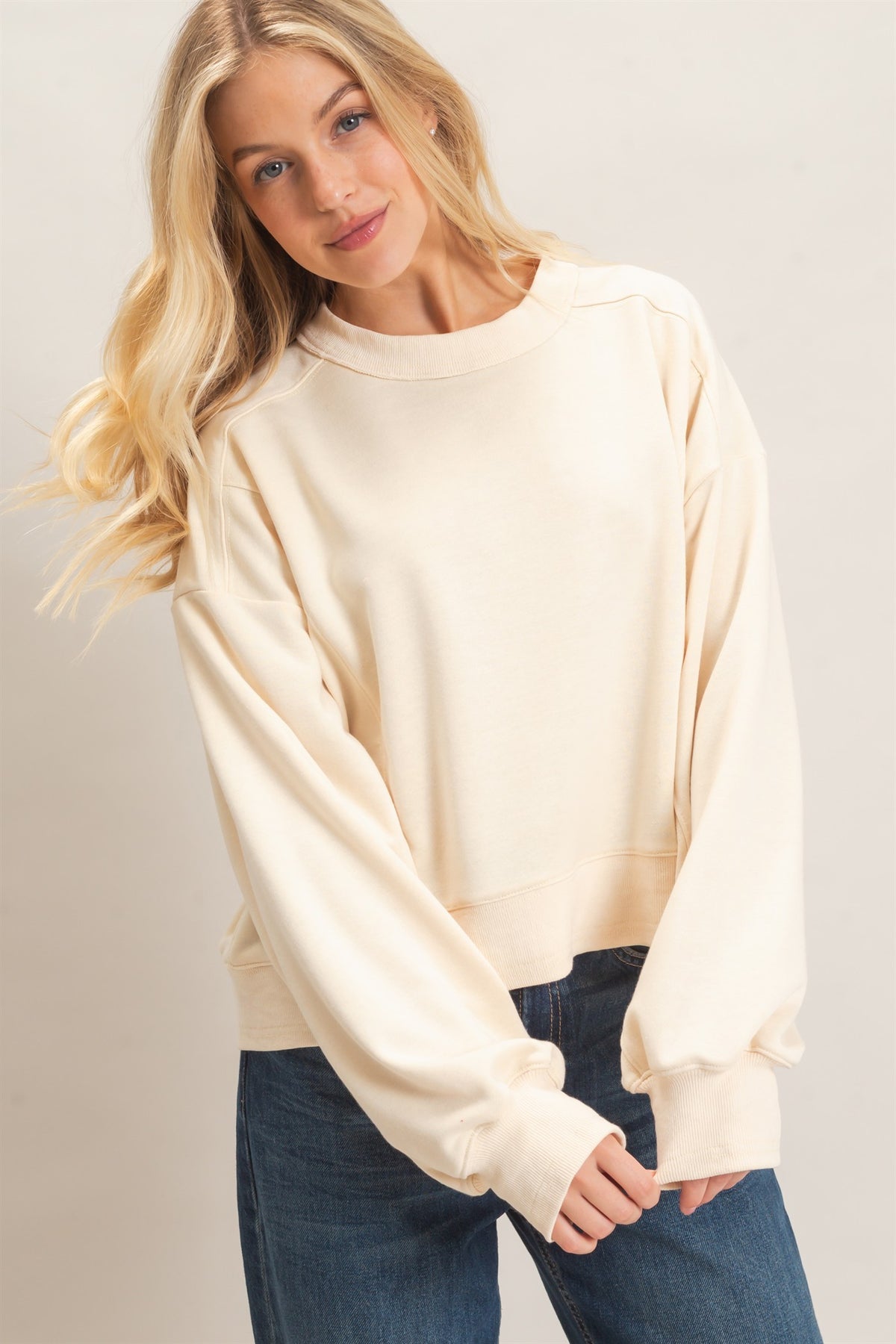 A woman wearing a cream-colored, long-sleeve crewneck sweatshirt and dark blue jeans, photographed against a neutral light gray studio background.