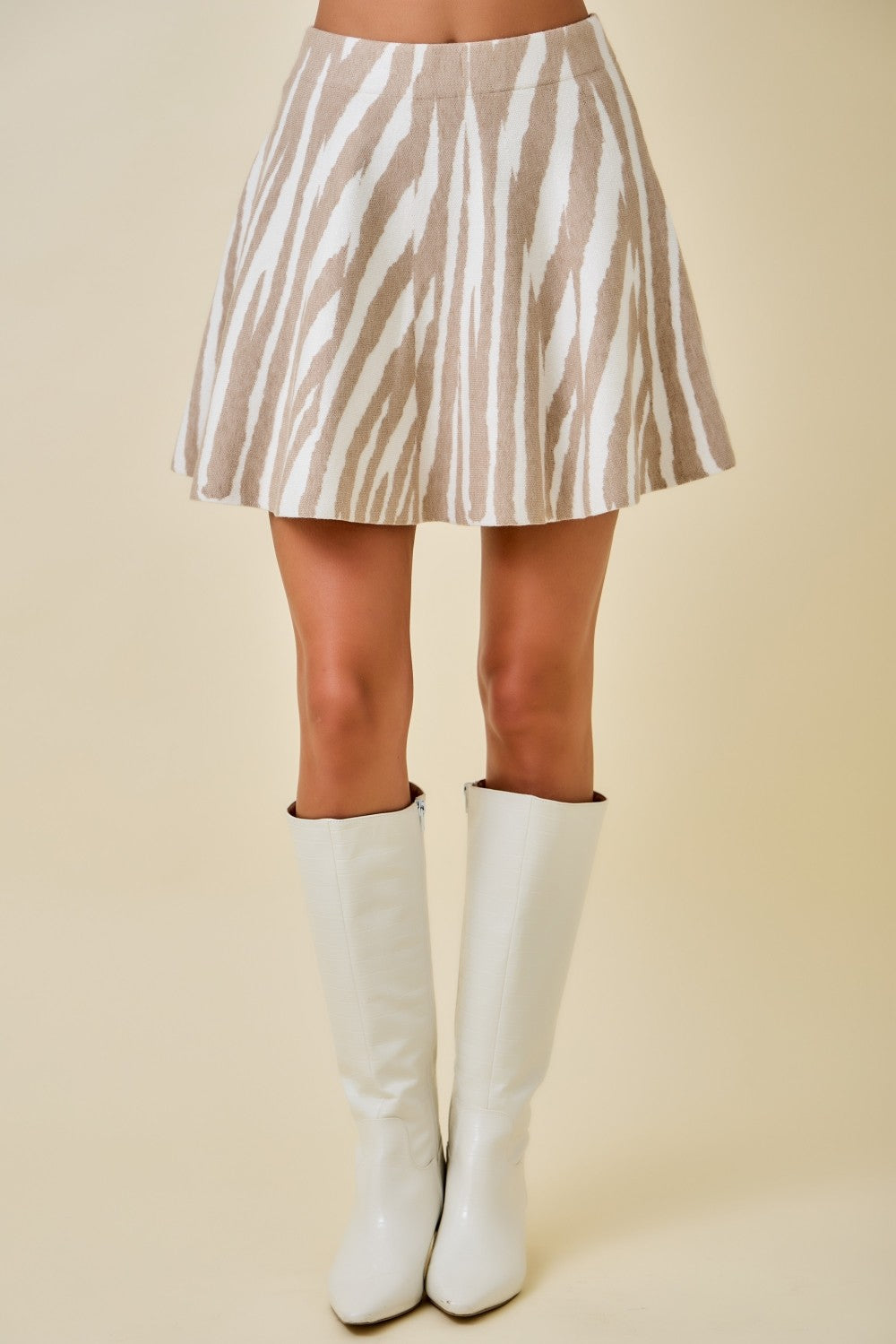 A model's lower body wearing a beige and white zebra-print mini skirt with white knee-high boots, set against a solid beige studio background.
