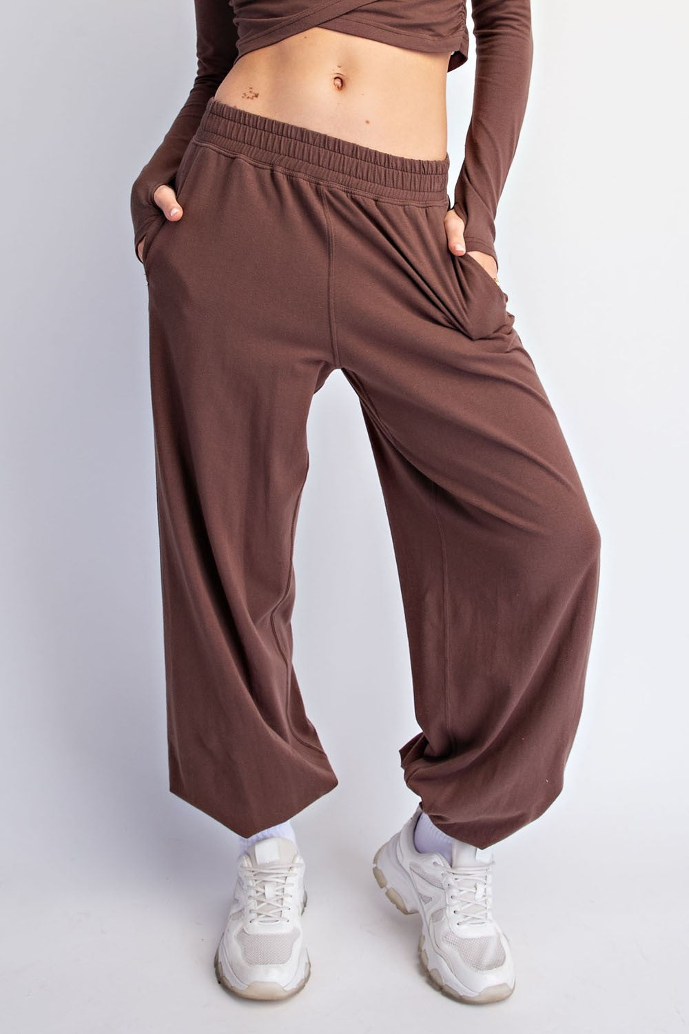 A person wearing loose, wide-leg brown pants with an elastic waistband and side pockets, paired with white sneakers, shown from the torso down against a light gray studio background.