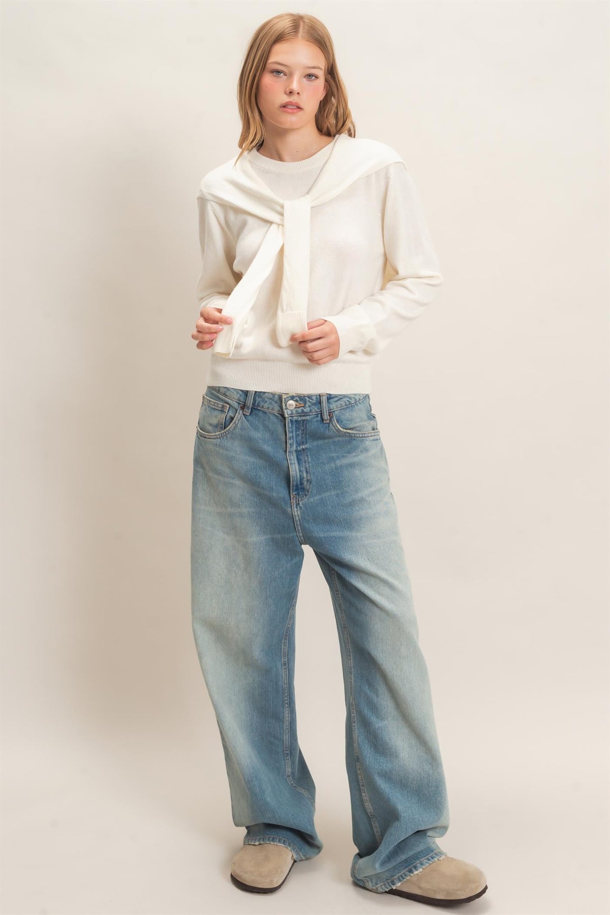 woman standing on white backdrop wearing cream sweater and jeans 