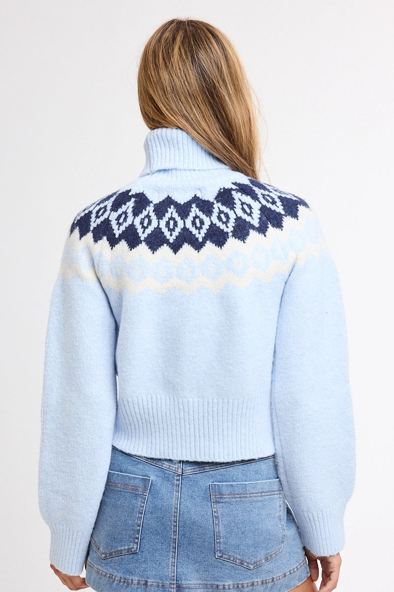 A woman viewed from the back wearing a light blue knit turtleneck sweater with a navy and cream geometric pattern across the shoulders, paired with blue denim shorts against a plain white background.